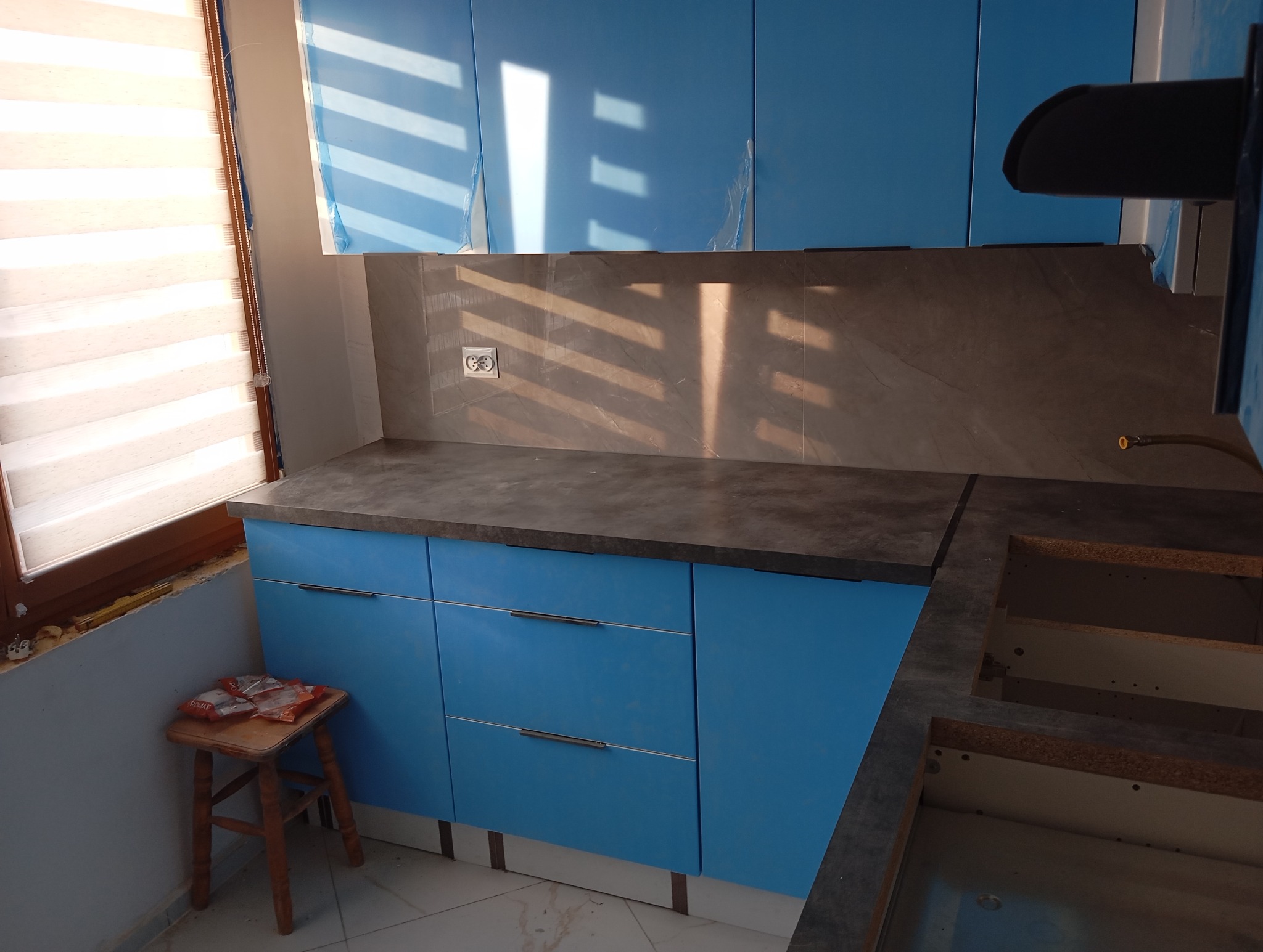 Niebieskie szafki kuchenne z szarym blatem i płytkami na ścianie, częściowo zabezpieczone folią ochronną, widoczny fragment okna z żaluzjami i taboret z akcesoriami montażowymi.