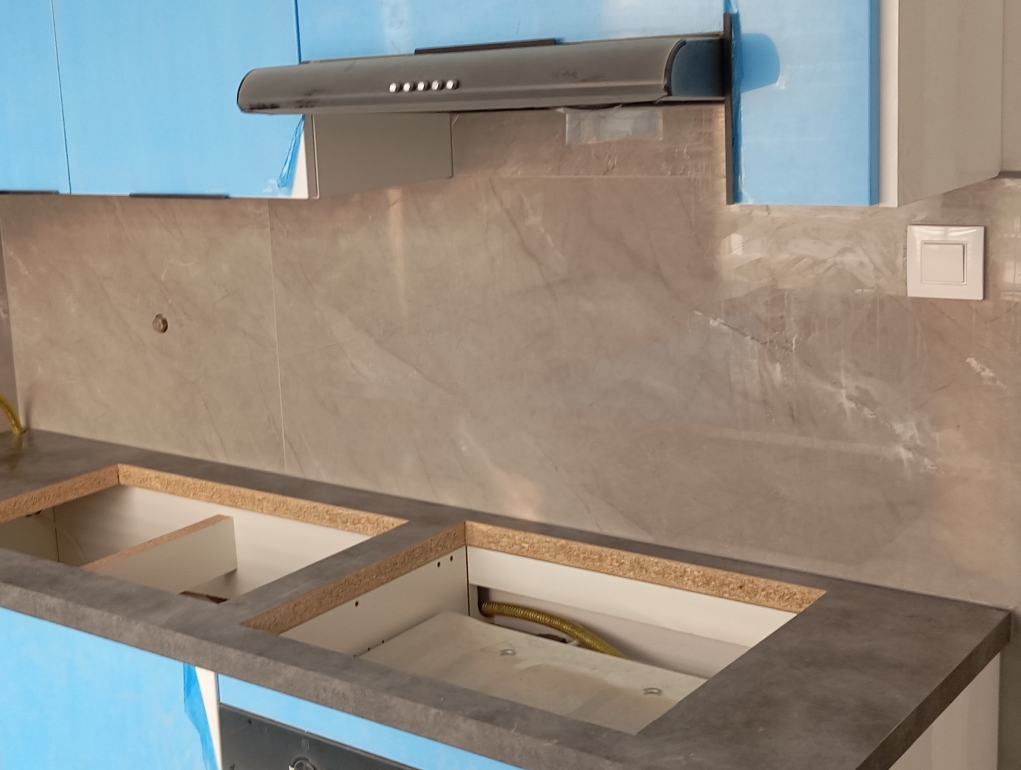 Niedokończony montaż szafek kuchennych z szarym blatem i marmurową ścianą, widoczne zabezpieczenia folią na niebieskich frontach, wyciąg kuchenny z oświetleniem.