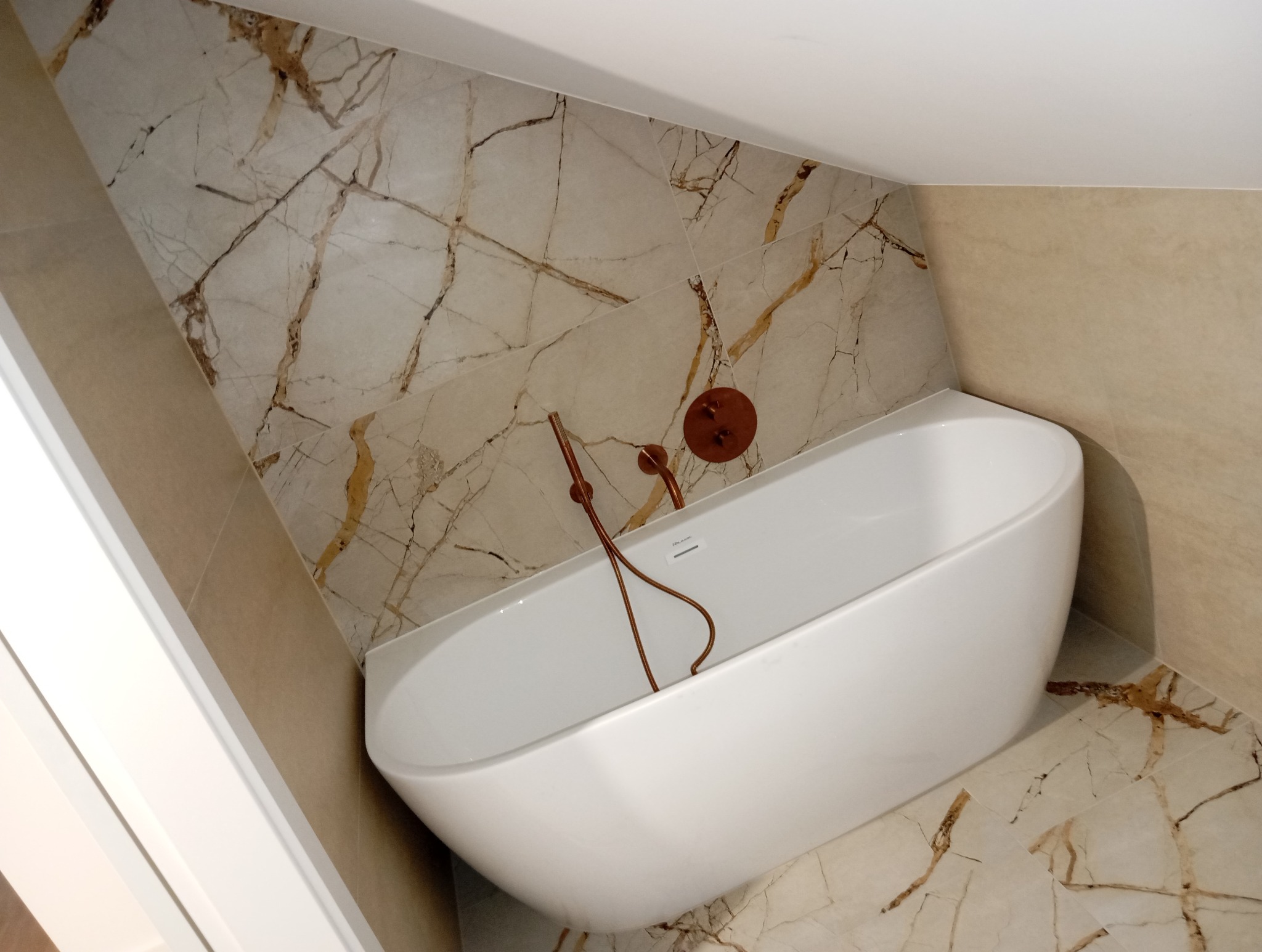 Nowoczesna biała wanna wolnostojąca z miedzianą armaturą na tle płytek imitujących marmur w łazience na poddaszu.