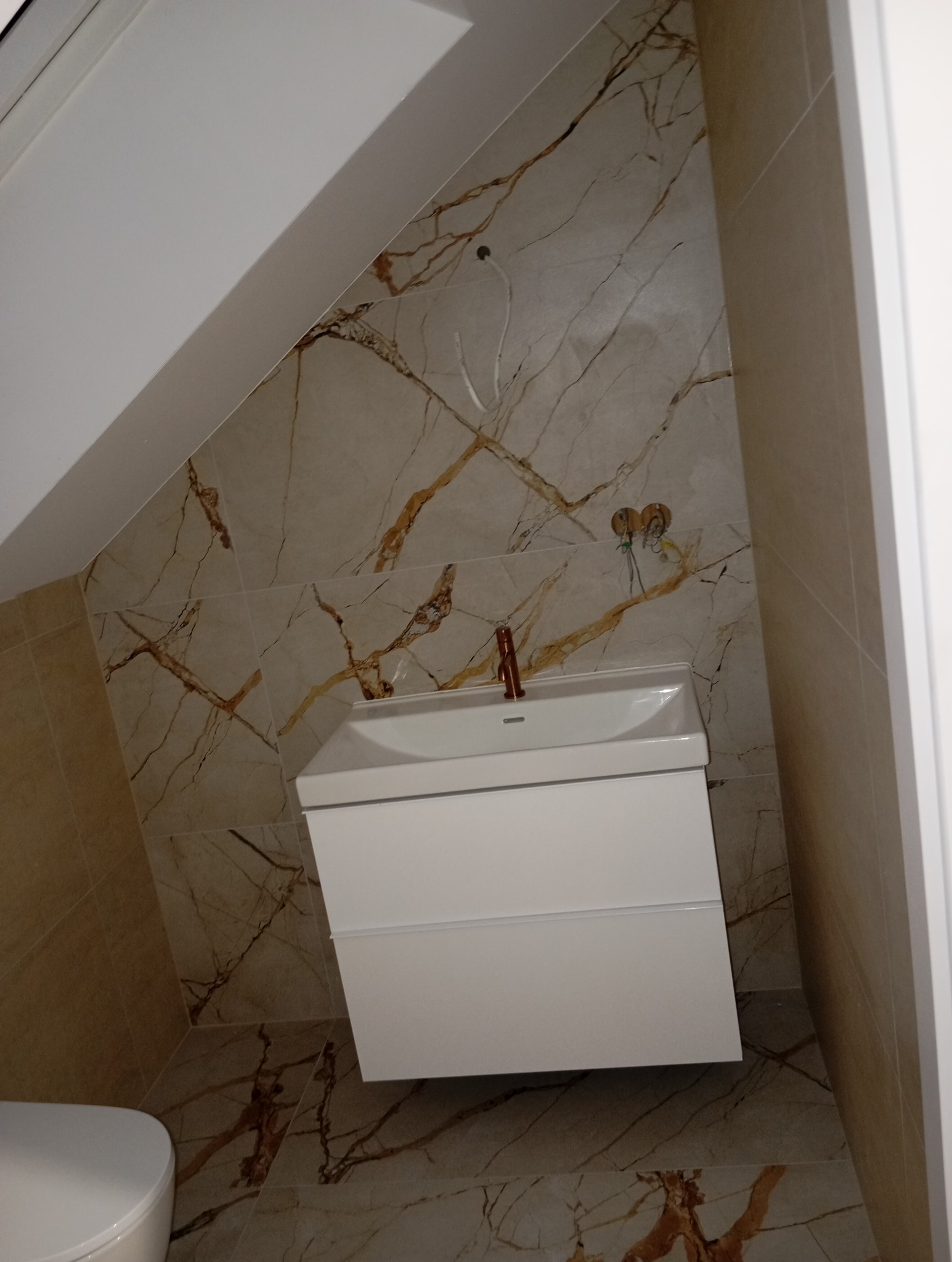 Ukośna łazienka na poddaszu z marmurowymi płytkami, białą szafką pod umywalkę i miedzianą baterią; widoczne niedokończone podłączenia elektryczne.