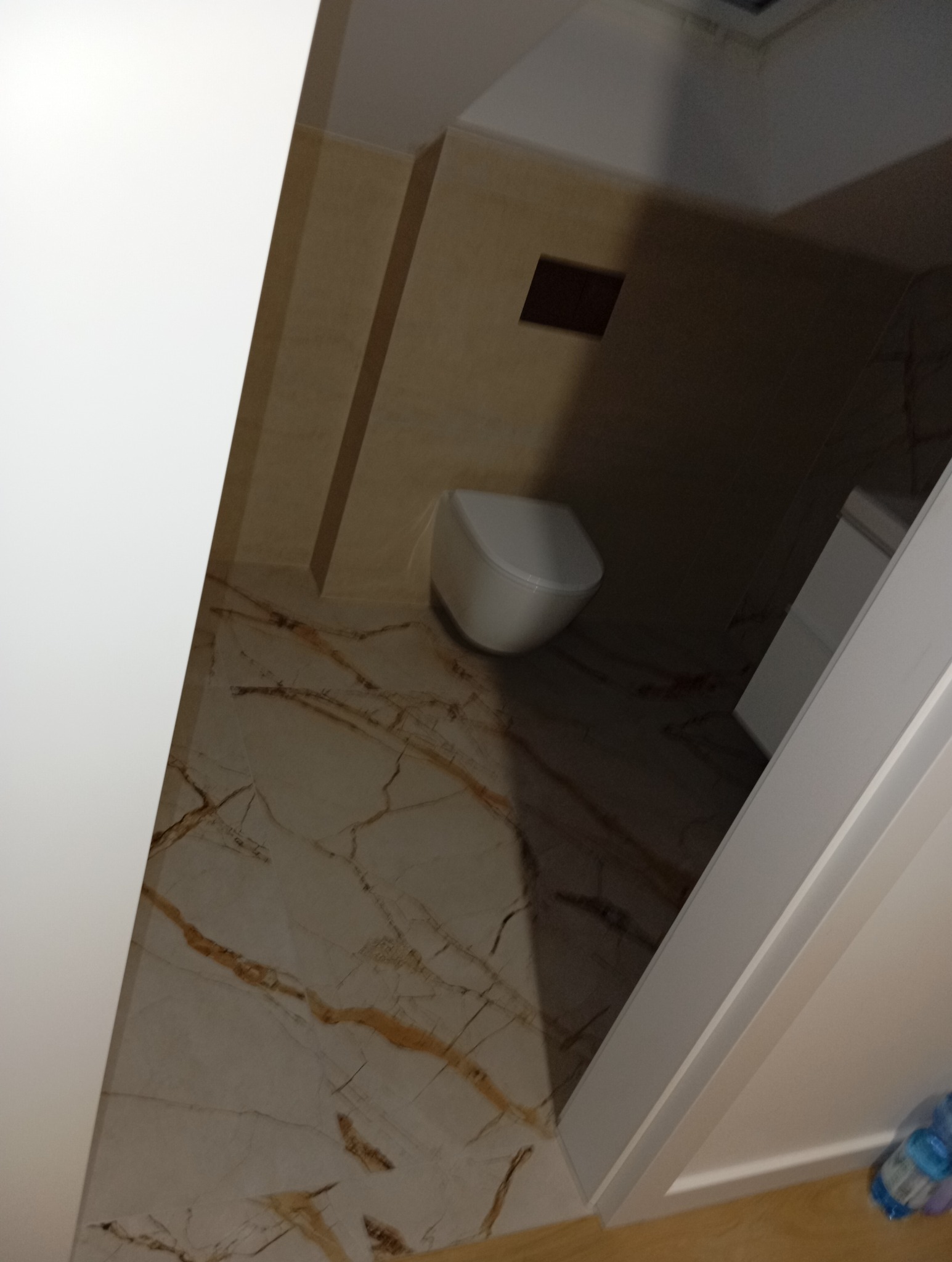 Wykończona łazienka z beżowymi płytkami imitującymi marmur, wisząca toaleta i szafka pod umywalką, widziana pod kątem z perspektywy drzwi.