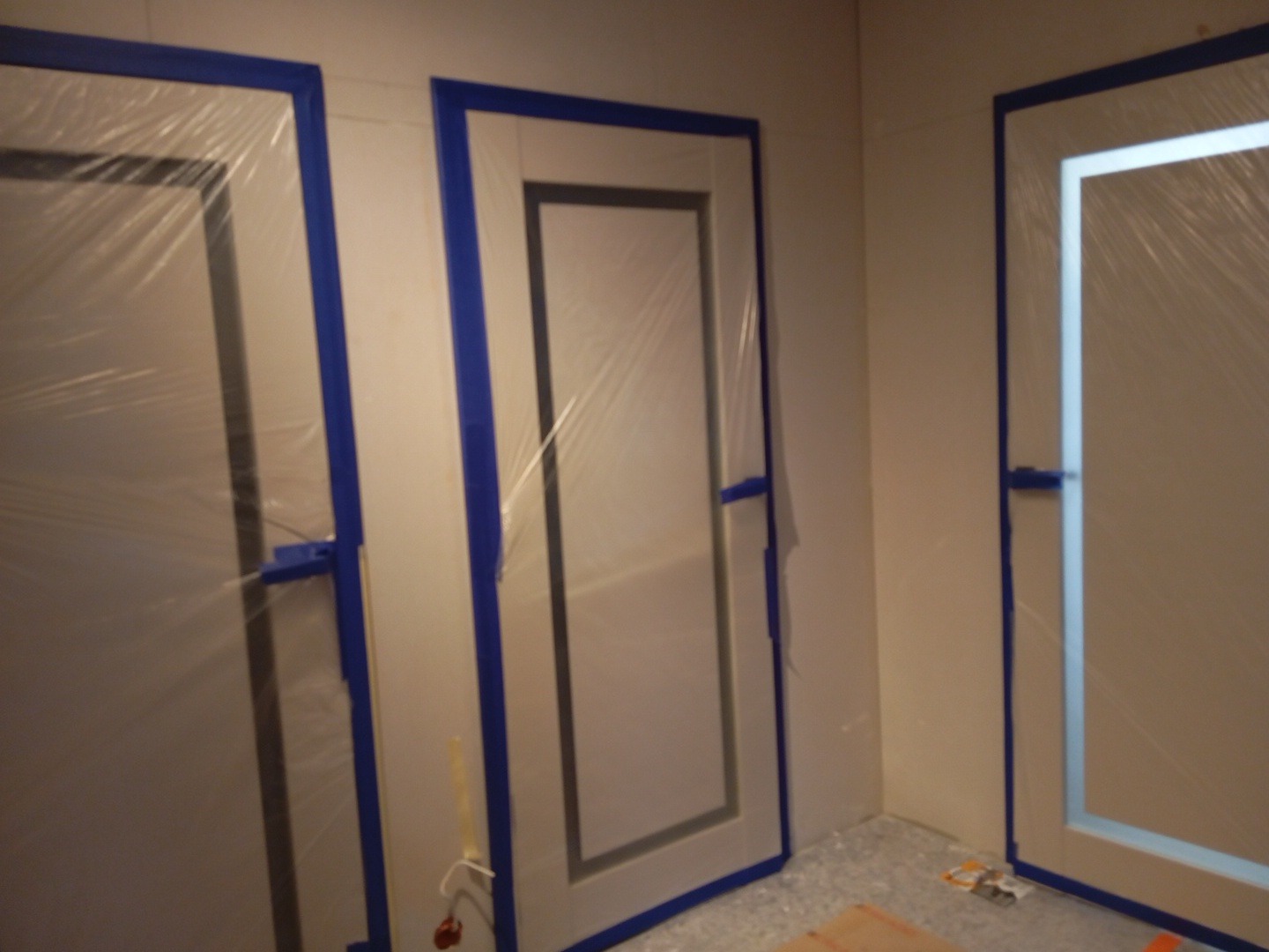 Przygotowanie do malowania: trzy drzwi zabezpieczone folią i niebieską taśmą malarską w rogu pokoju.