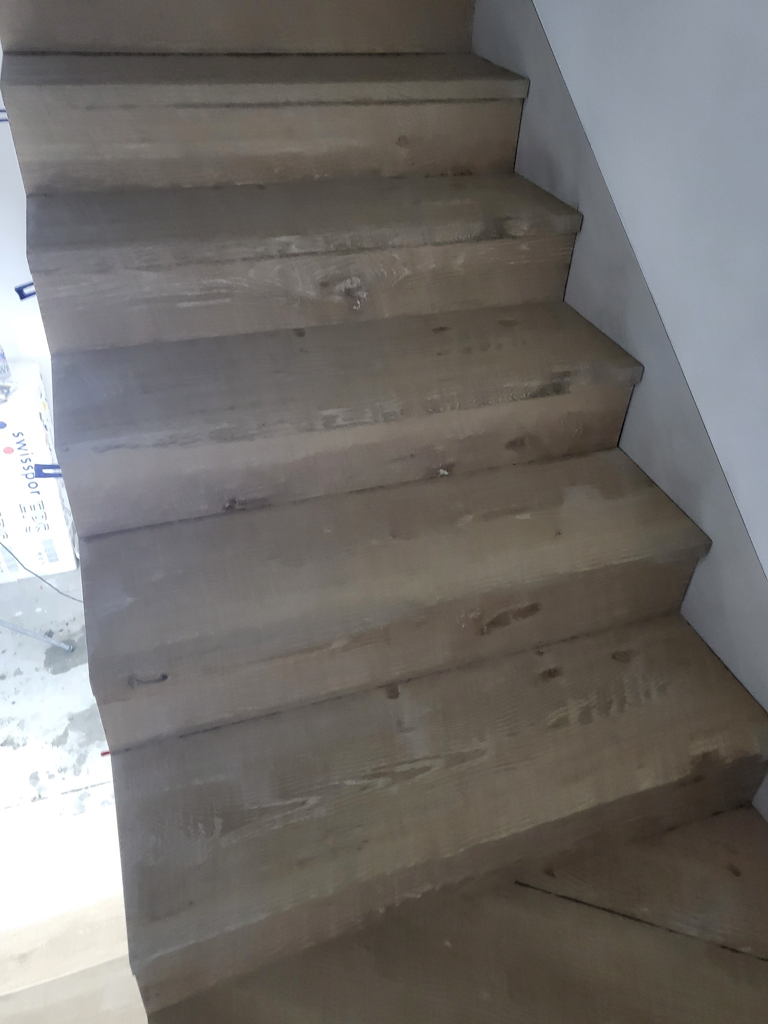 Drewniane schody o jasnym, naturalnym odcieniu, widoczne z dołu, z lekkim zabrudzeniem po remoncie i resztkami folii ochronnej w rogu.