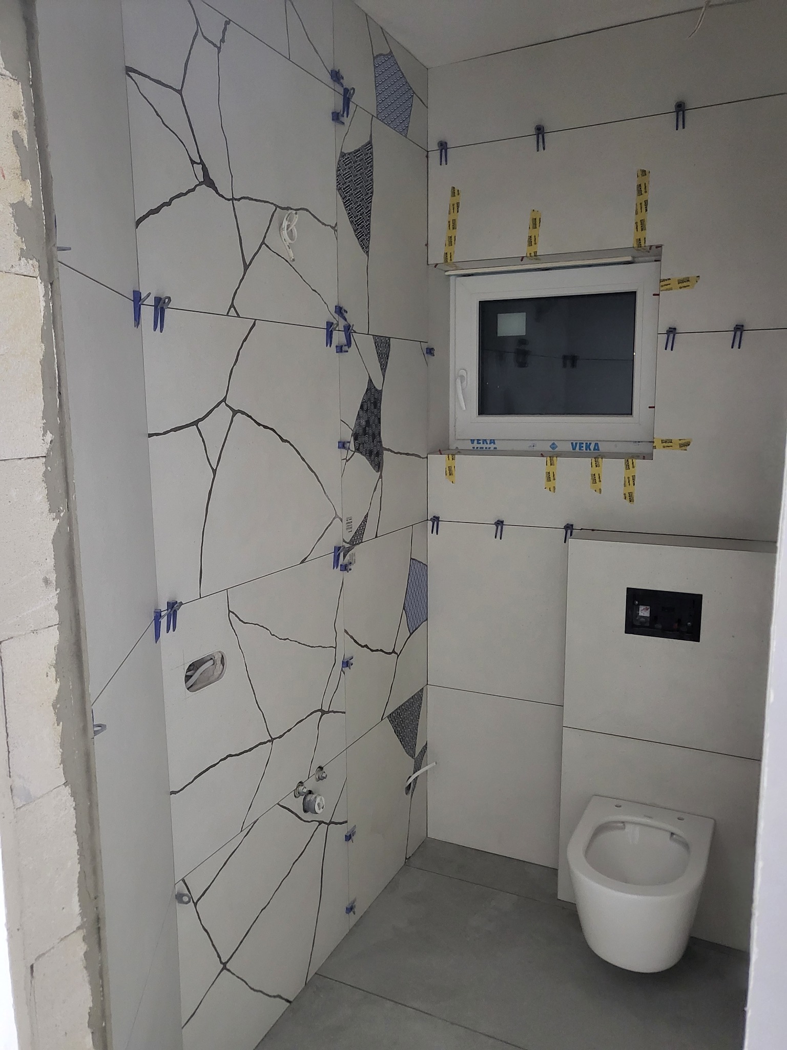 Białe płytki imitujące pęknięcia na ścianie łazienki w trakcie montażu, widoczne klipsy poziomujące, okno z naklejoną taśmą, podwieszana toaleta, surowy beton na podłodze.