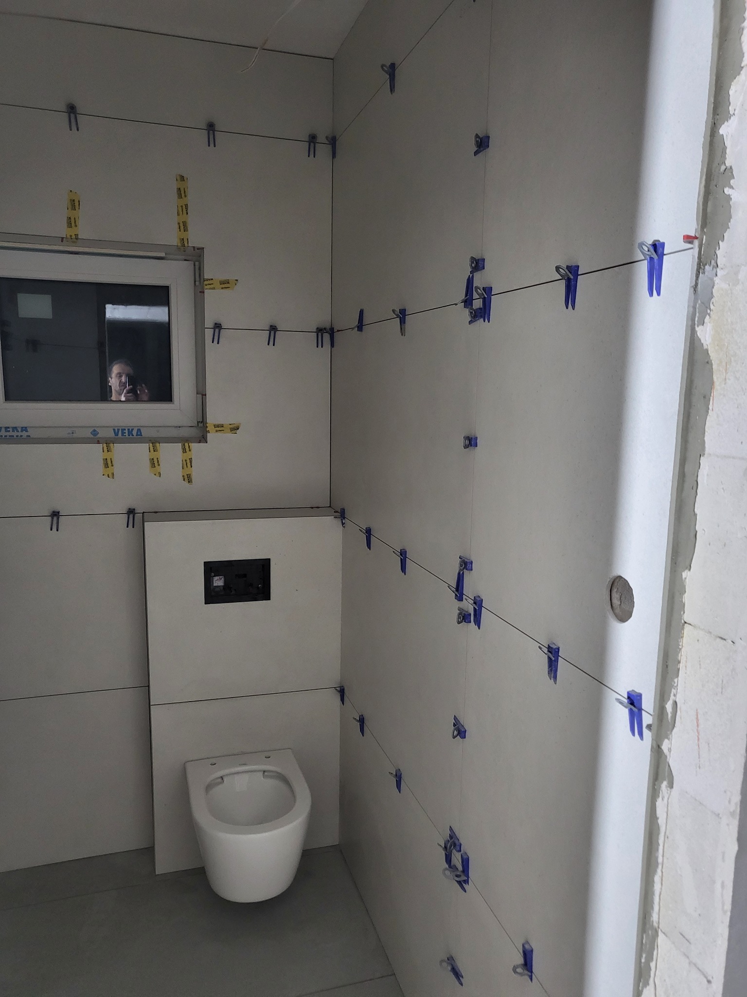 Świeżo ułożone, duże, jasnoszare płytki na ścianach łazienki, utrzymywane w poziomie przez niebieskie klipsy i żyłki; w tle widoczne okno z odbiciem osoby robiącej zdjęcie, pod oknem zabudowa ze...