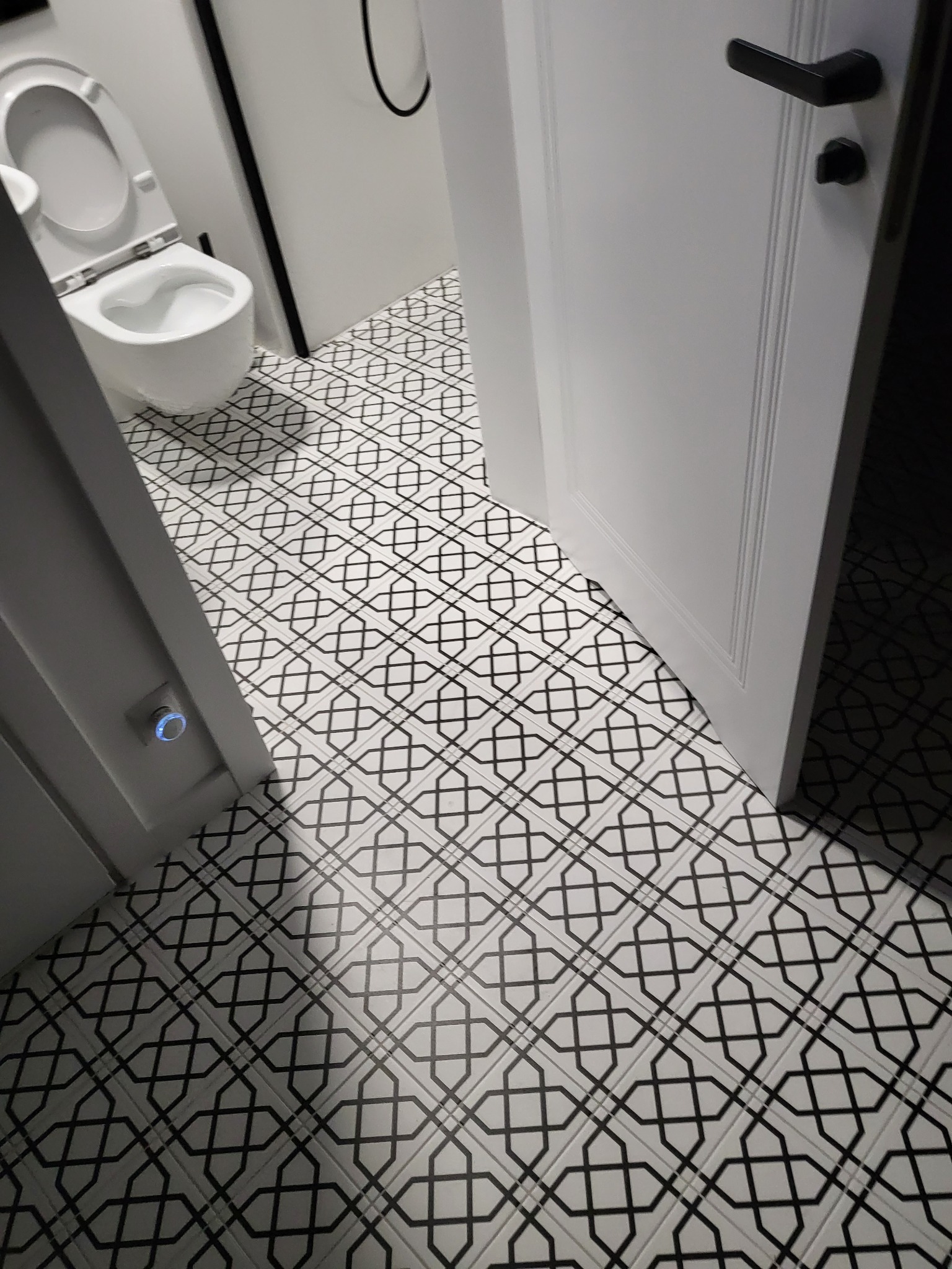 Biała łazienka z toaletą i geometryczną podłogą z płytek, widok zza uchylonych drzwi.