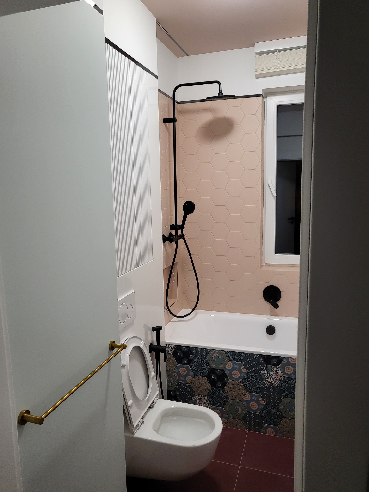 Nowoczesna łazienka z różowymi heksagonalnymi płytkami nad wanną i czarną armaturą, widoczna otwarta toaleta z uchwytem na drzwiach.