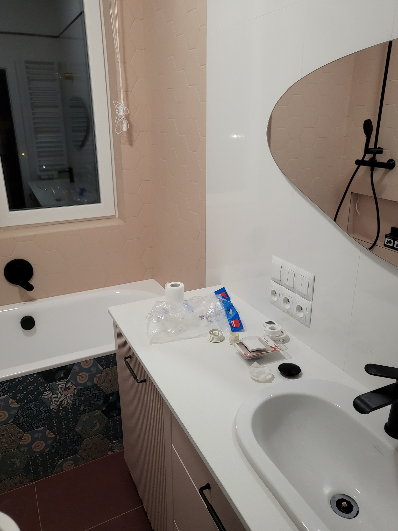 Łazienka po remoncie z heksagonalnymi płytkami w odcieniach różu na ścianach, białą armaturą i czarną baterią umywalkową, widoczne akcesoria montażowe na blacie.