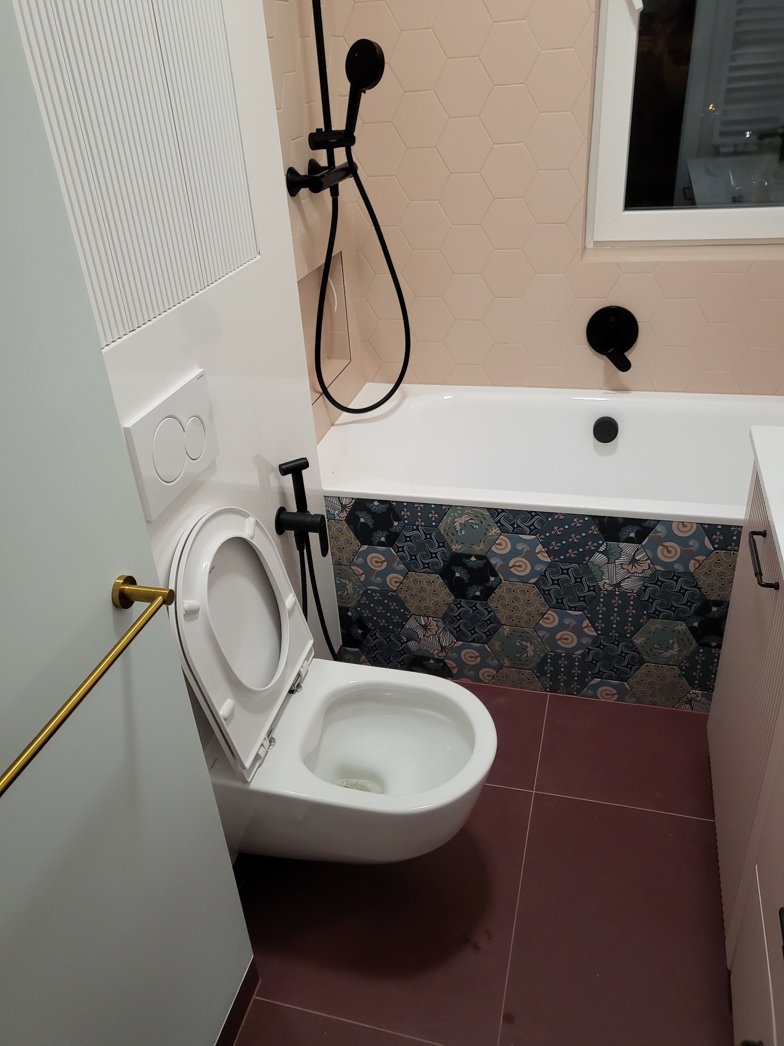 Nowoczesna łazienka z heksagonalnymi płytkami w odcieniach beżu i granatu, czarna armatura, biała wanna i toaleta z otwartą deską, brązowe płytki podłogowe.