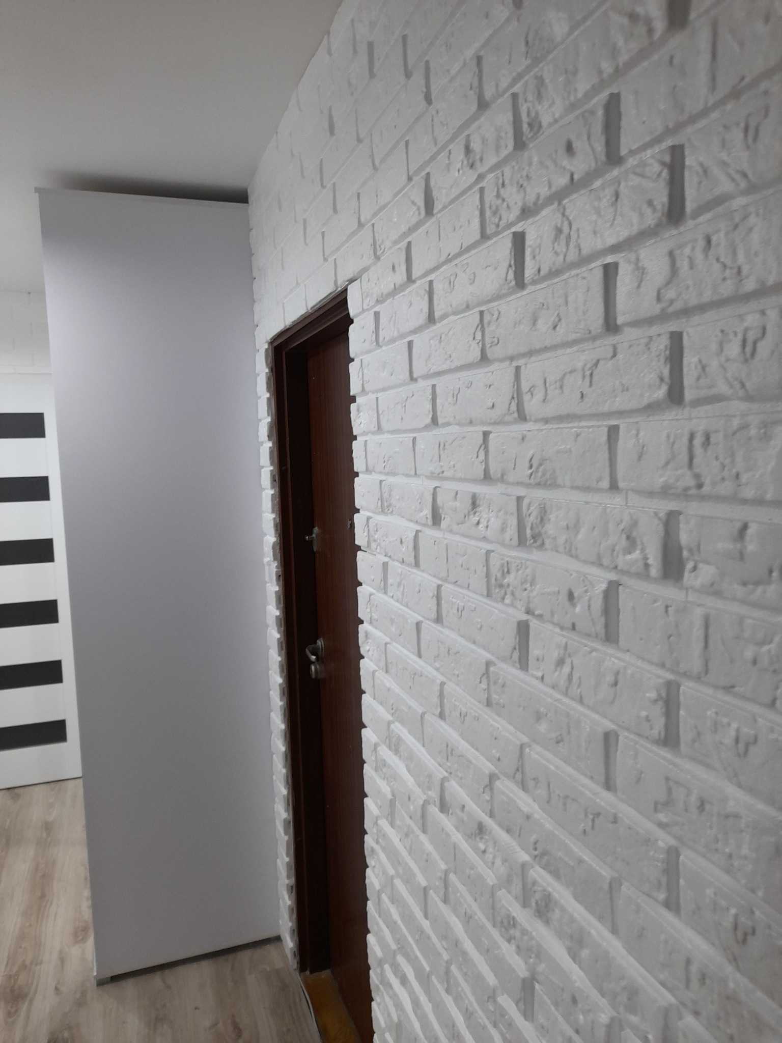 Ściana z białej cegły dekoracyjnej z brązowymi drzwiami i fragmentem drugiej ściany w tle.