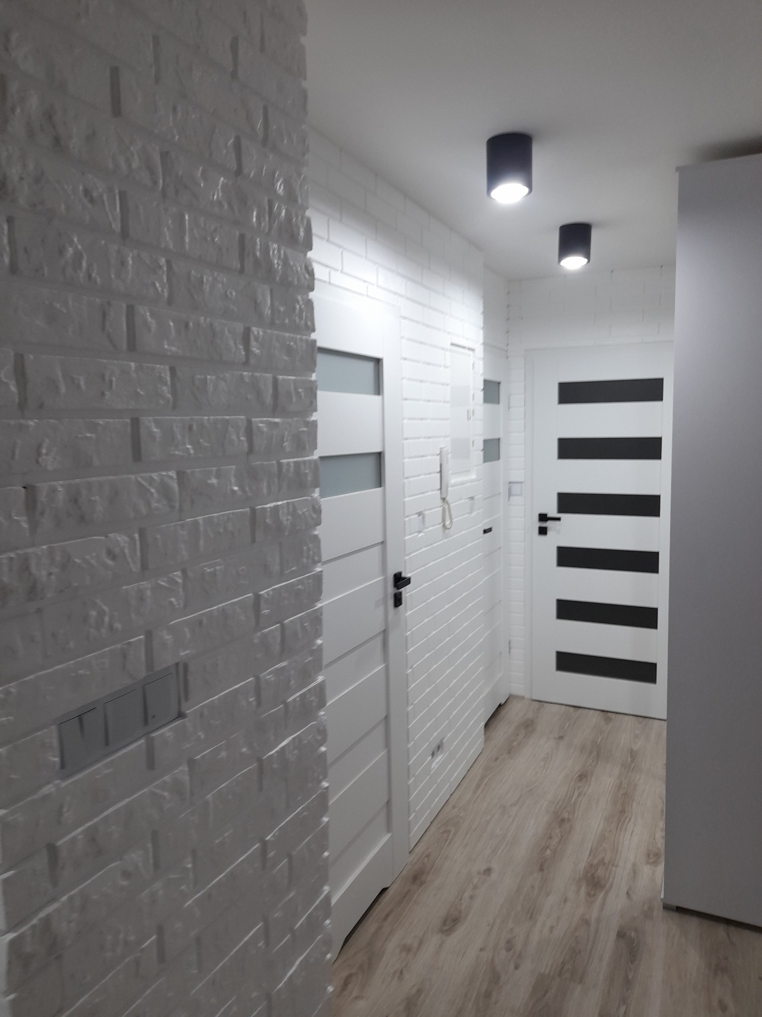 Korytarz wykończony białą cegłą, z dwiema białymi drzwiami z czarnymi akcentami i drewnopodobną podłogą, oświetlony czarnymi lampami sufitowymi.