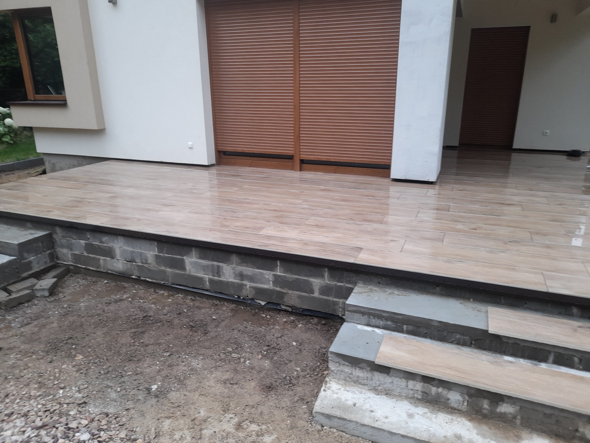 Wykończony taras z brązowych płytek imitujących drewno, schody w trakcie budowy, widoczne niedokończone elementy i surowy grunt.