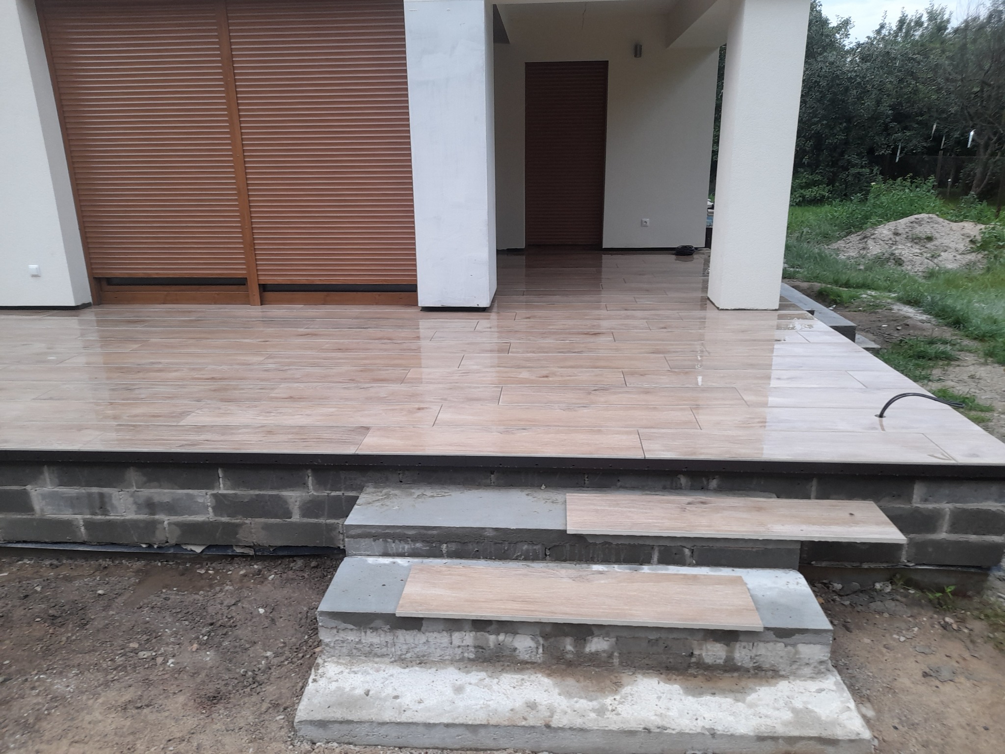 Układanie płytek imitujących drewno na tarasie z trzema schodami, widoczne niedokończone fugowanie i świeżo ułożone płytki na schodach.