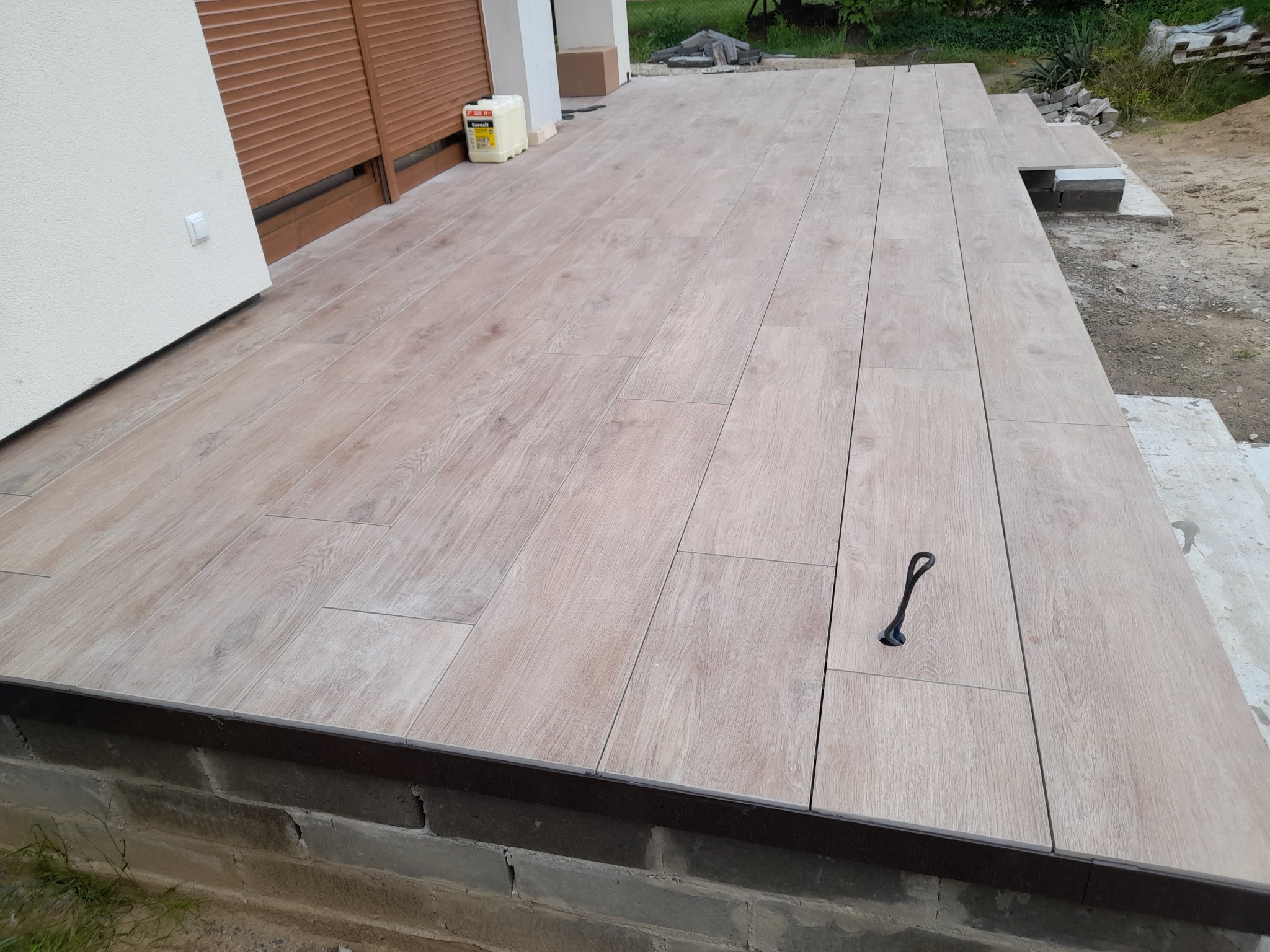 Wyłożony płytkami taras imitującymi drewno, z widocznym fragmentem elewacji budynku i pojemnikiem z klejem na boku.