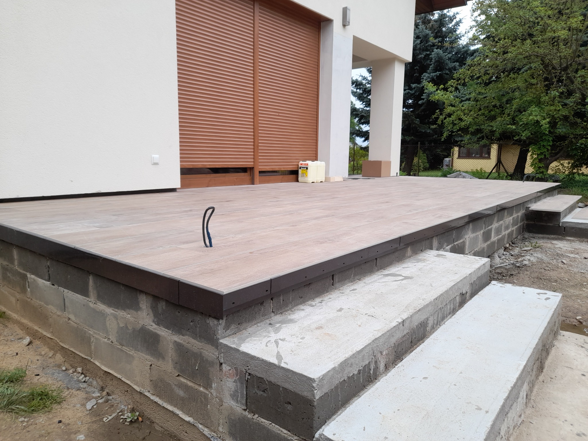 Wyłożony płytkami taras z betonowymi schodami, z widocznym pojemnikiem z klejem do płytek i brązowymi roletami okiennymi w tle.