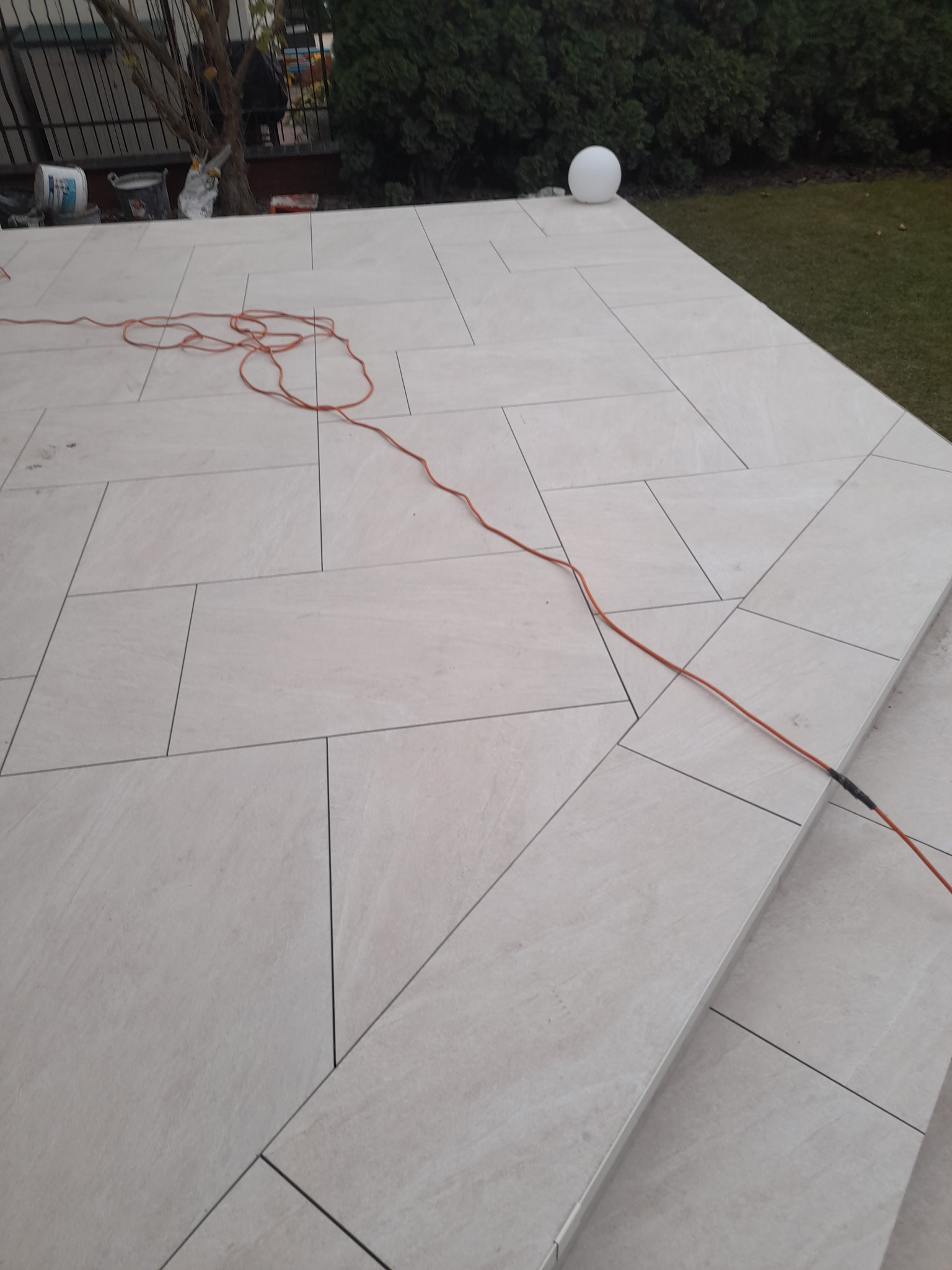 Ułożona, jasna, prostokątna terakota na zewnątrz, widoczny pomarańczowy kabel zasilający, lampa-kula i roślinność w tle.