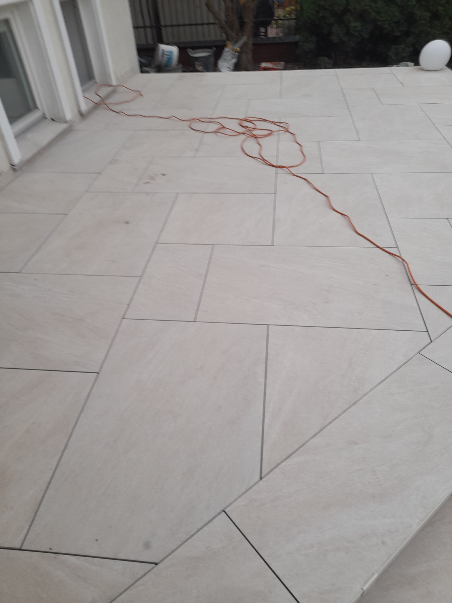 Jasne, prostokątne płytki ułożone na tarasie, z pomarańczowym przewodem elektrycznym leżącym na powierzchni, widoczne okno i biała kula dekoracyjna w tle.
