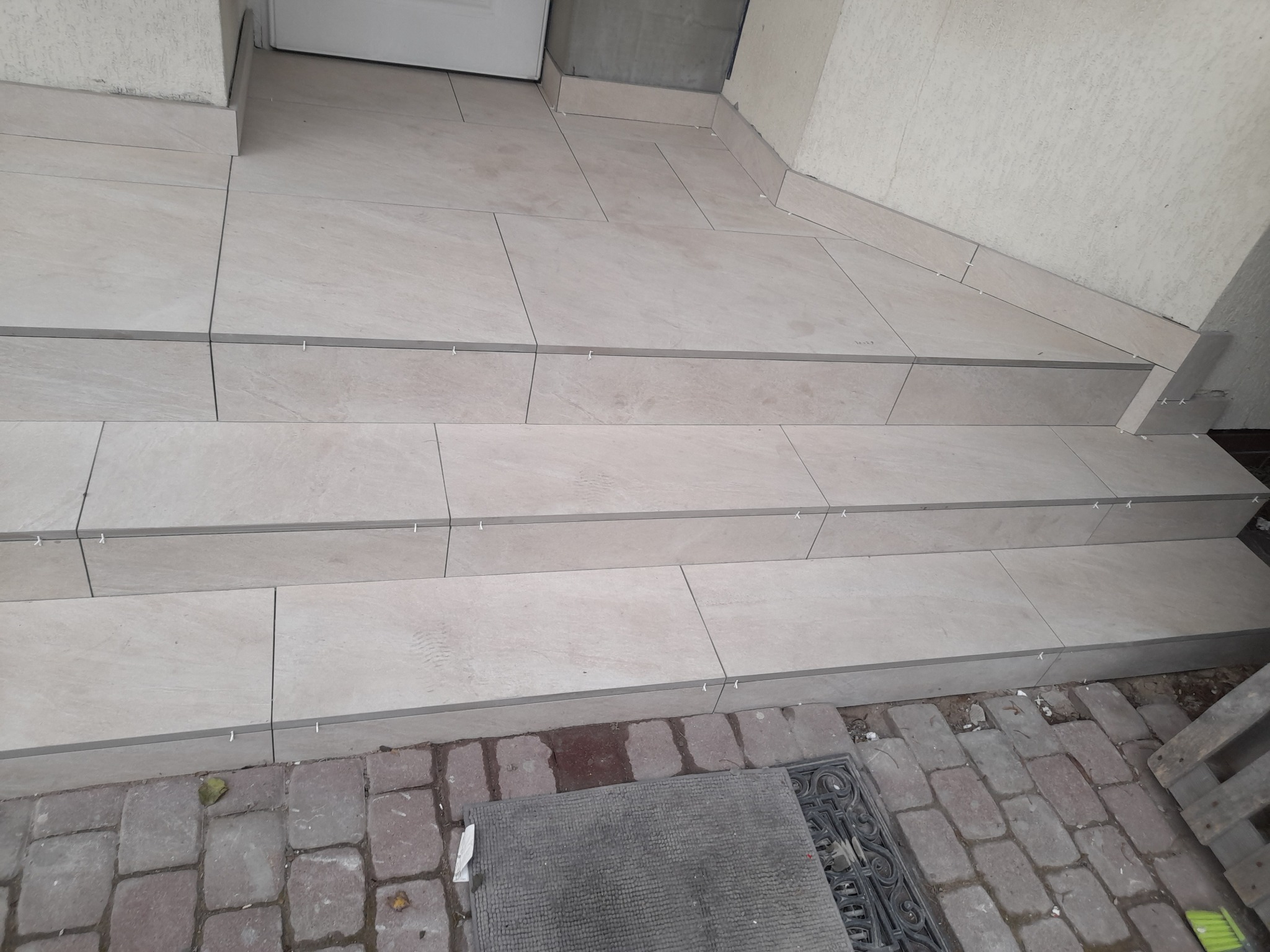 Wykończone schody zewnętrzne z jasnych, prostokątnych płytek ceramicznych z widocznymi krzyżykami dystansowymi, prowadzące do drzwi wejściowych, z fragmentem kostki brukowej i wycieraczki u podstawy.