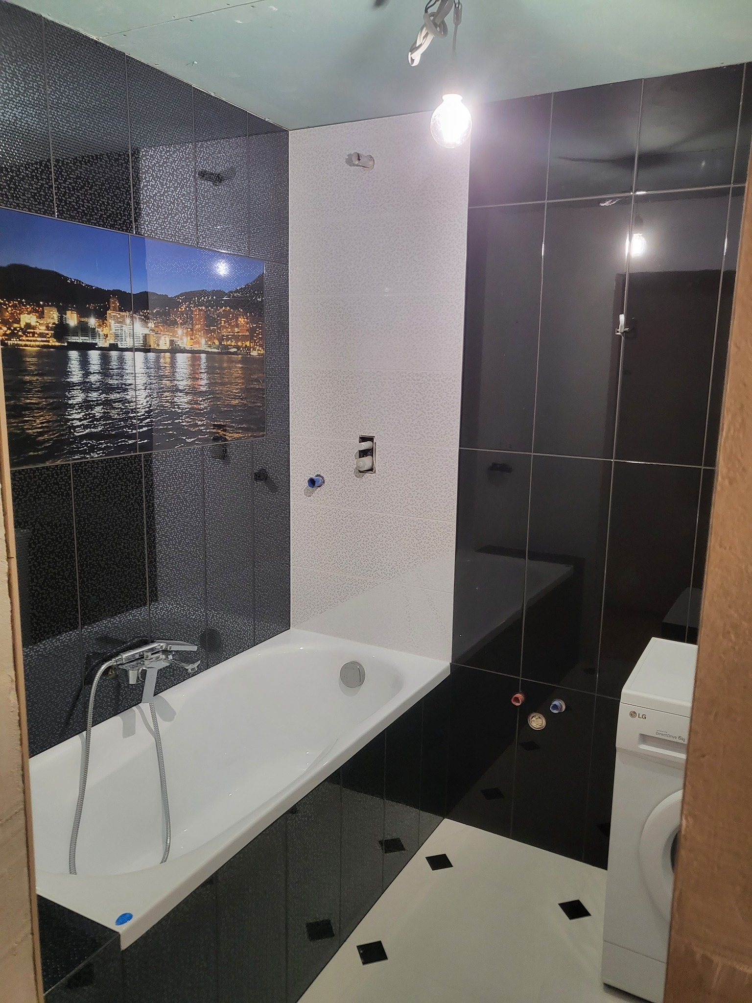 Wanna obudowana czarno-białymi płytkami, z baterią i odpływem, na ścianie płytki z motywem miasta nocą, obok pralka LG, widoczne niedokończone prace wykończeniowe.