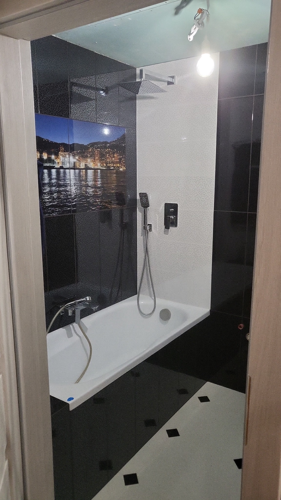 Wykończona łazienka z wanną, czarno-białymi płytkami i fototapetą z nocnym widokiem miasta nad wodą.