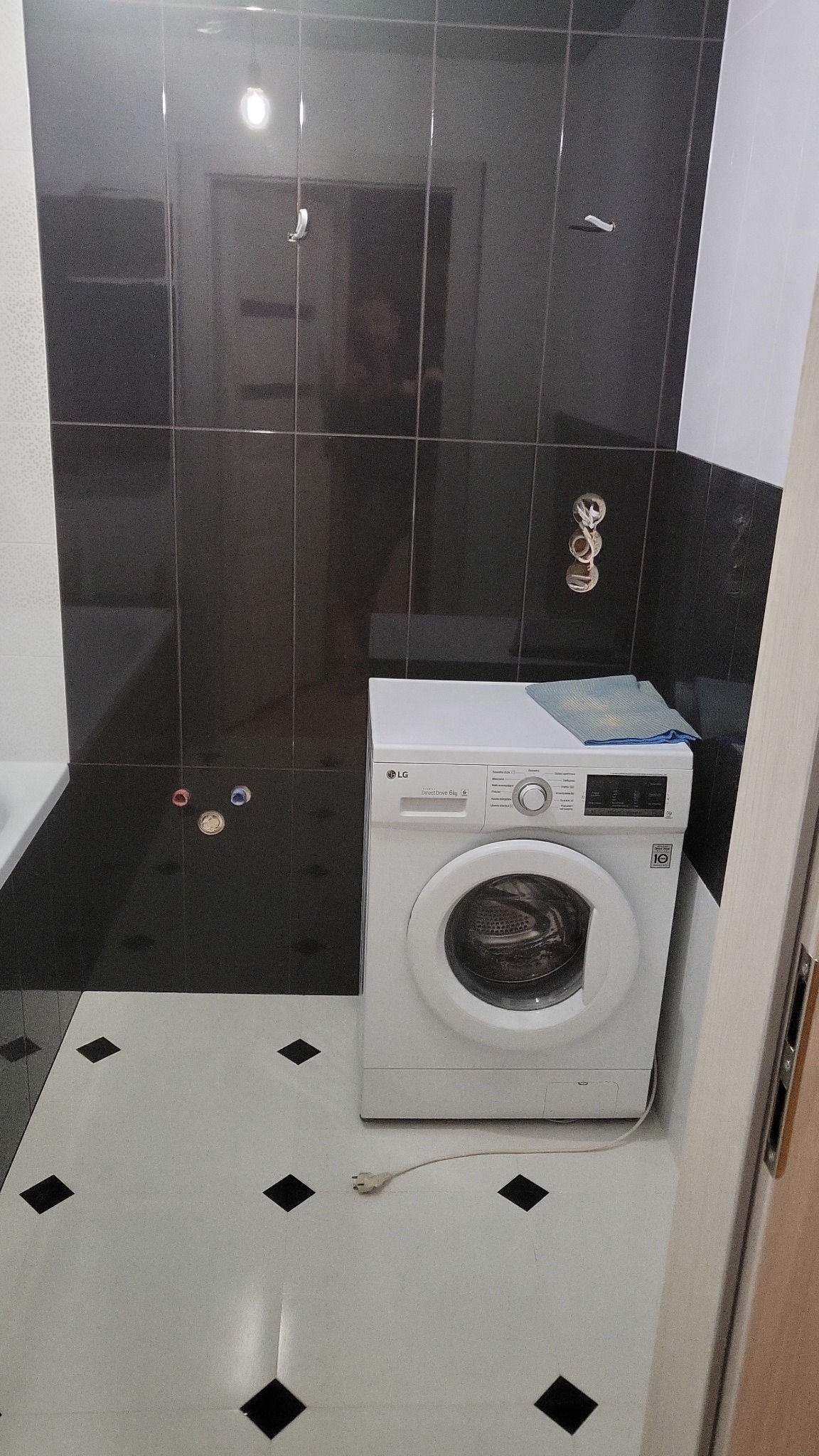 Łazienka z czarnymi płytkami ściennymi, białą podłogą w romby i pralką LG z niebieską ścierką na górze.