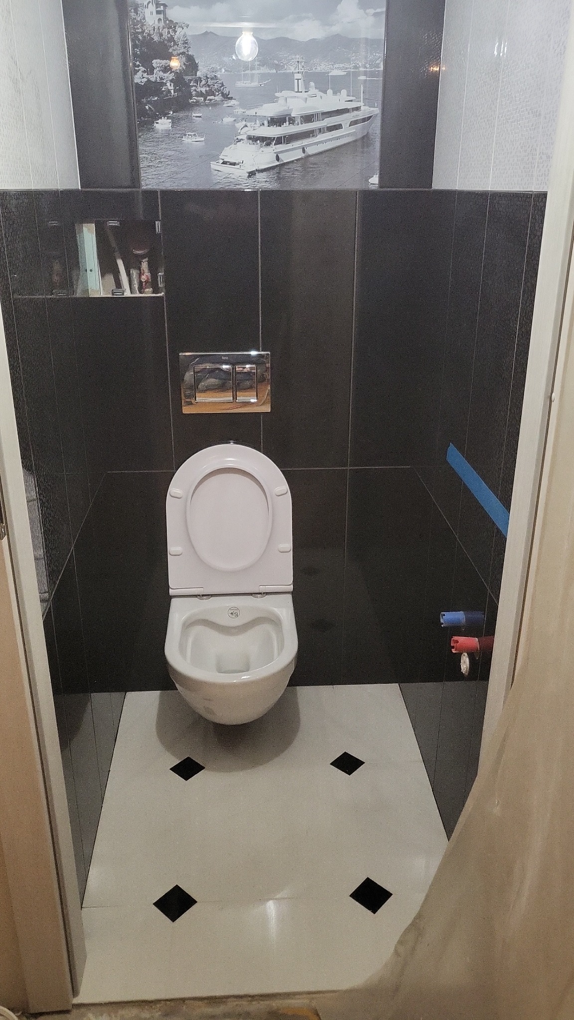 Toaleta z deską wolnoopadającą na podłodze z białych płytek z czarnymi rombami, otoczona czarnymi, błyszczącymi płytkami ściennymi, nad toaletą widoczny jest przycisk spłuczki oraz obraz z motywem...