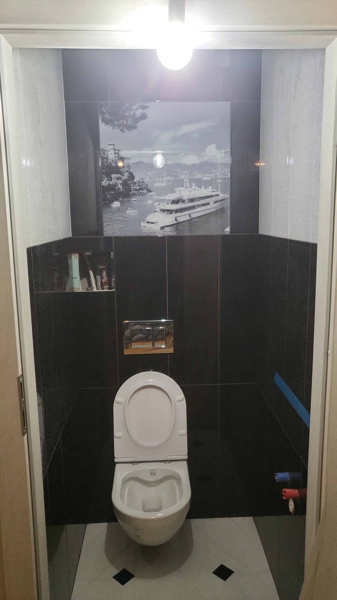 Nowoczesna toaleta w czarno-białej łazience z płytkami, nad nią grafika z jachtem, widoczny fragment niebieskiej taśmy malarskiej na ścianie.