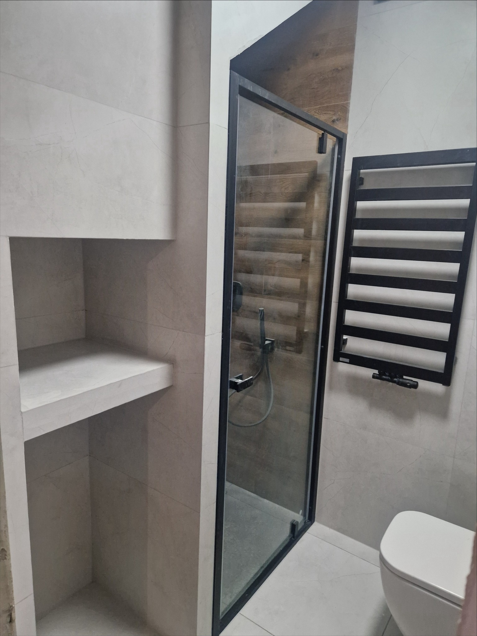 Nowoczesna łazienka z kabiną prysznicową w czarnej ramie, grzejnikiem drabinkowym i wnęką półkową wykończoną płytkami imitującymi marmur.