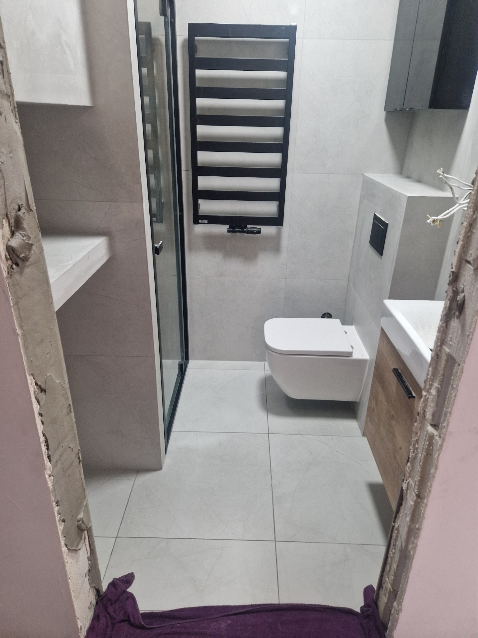 Nowoczesna łazienka z kabiną prysznicową, wiszącą toaletą i grzejnikiem drabinkowym, widziana z perspektywy niedokończonego wejścia z widocznymi śladami prac budowlanych i fioletowym ręcznikiem...