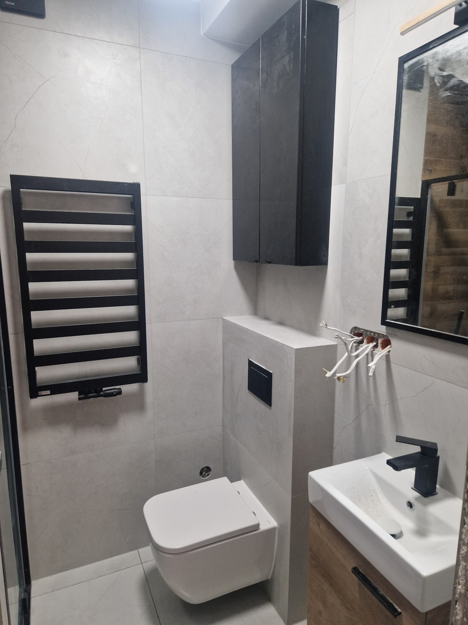 Nowoczesna łazienka w trakcie wykańczania: wisząca toaleta, czarny grzejnik drabinkowy, szafka z umywalką i czarną armaturą, oraz czarna szafka wisząca, z widocznymi przewodami elektrycznymi...