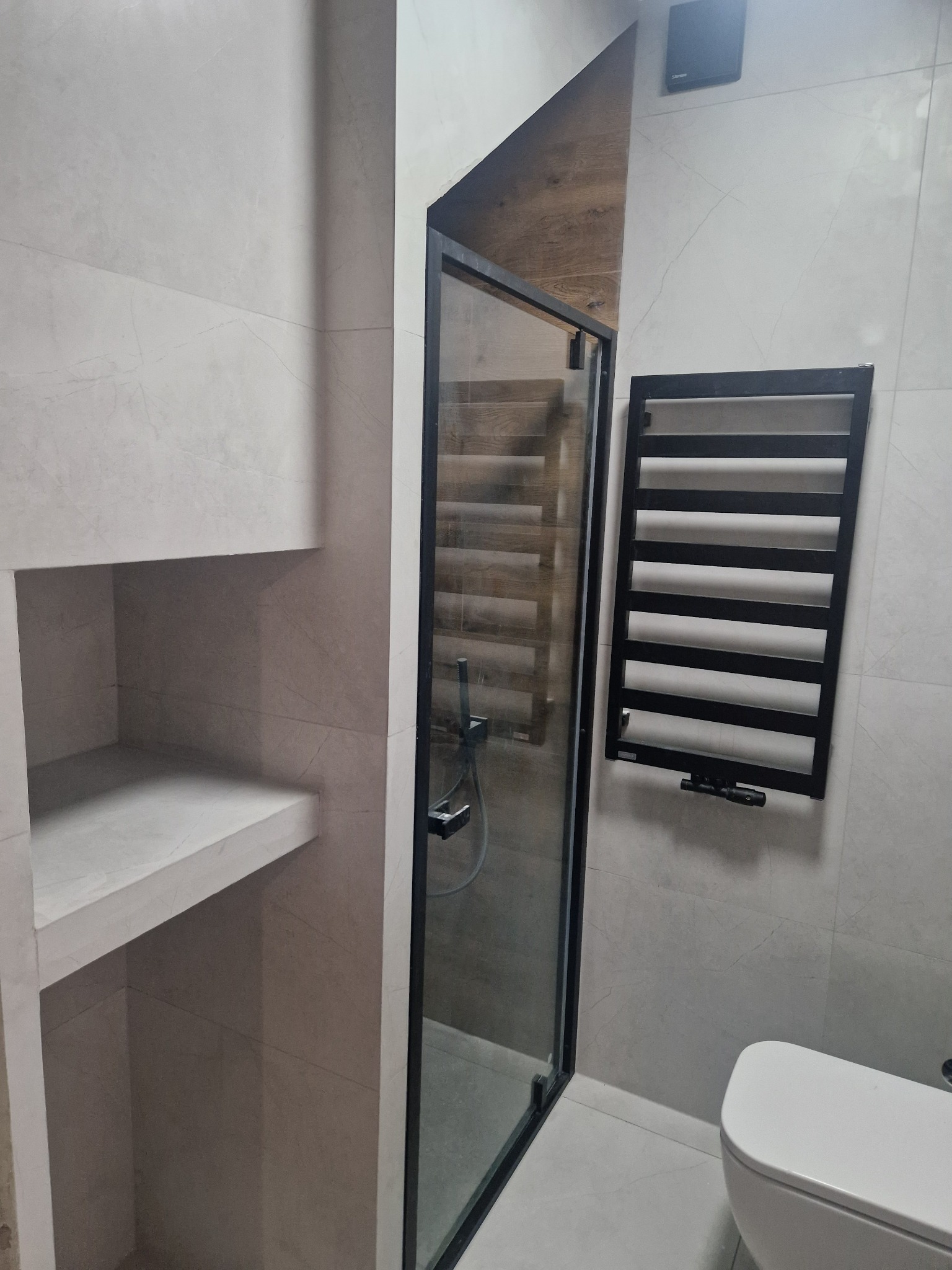 Nowoczesna łazienka z kabiną prysznicową z czarną ramą, grzejnikiem drabinkowym w czarnym kolorze i wnęką z półkami, wykończona płytkami w odcieniach szarości.