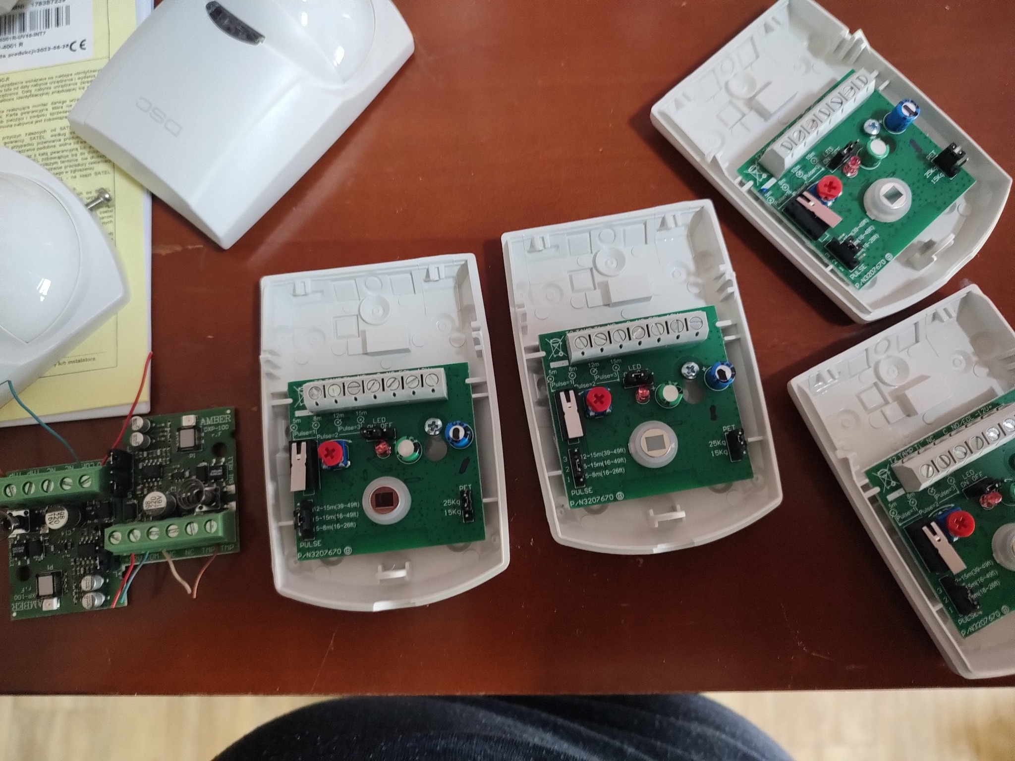 Rozłożone czujki alarmowe DSC oraz płytki elektroniczne AMBER na brązowym blacie, widoczne elementy montażowe i ustawienia czułości