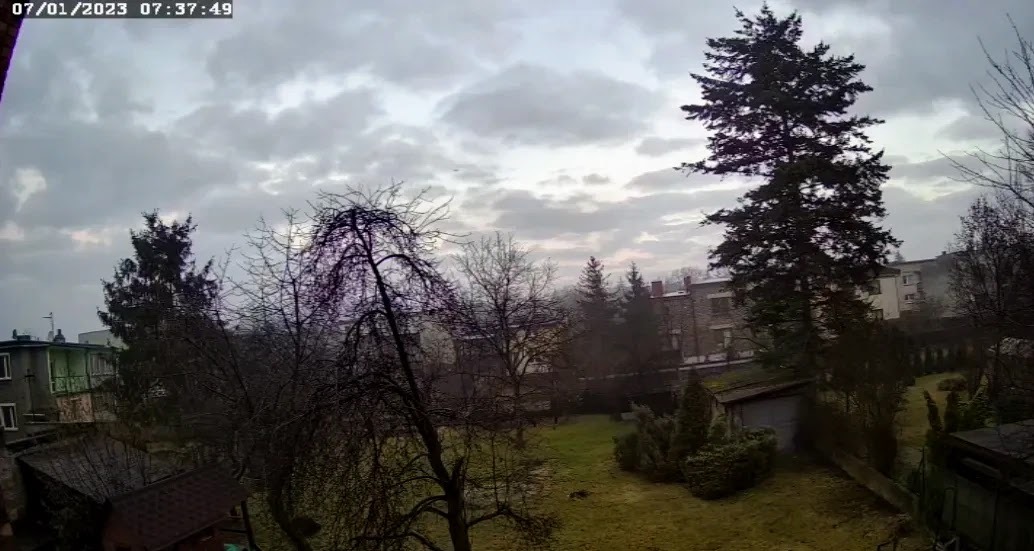 Kadr z kamery przemysłowej: widok na podwórze z budynkami w tle, pochmurne niebo, data i godzina w lewym górnym rogu.