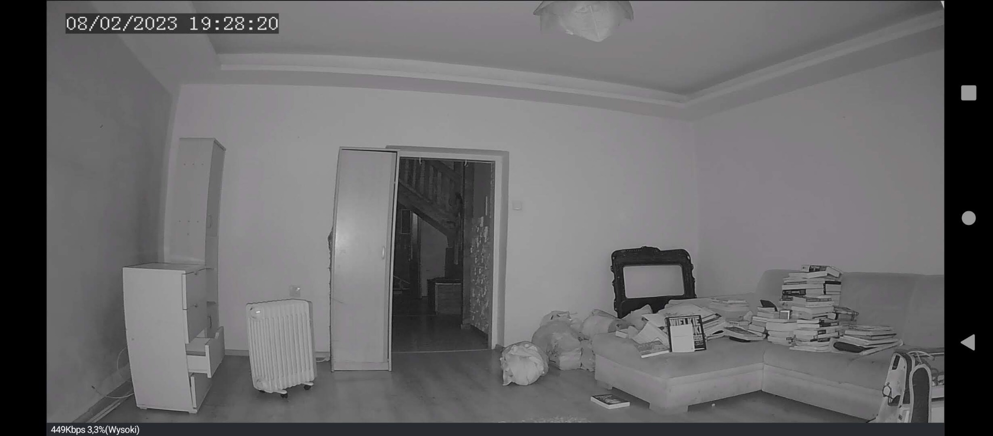 Czarno-biały obraz z kamery IP przedstawiający wnętrze pokoju z widocznym oświetleniem sufitowym, meblami, stertą książek na kanapie i datą w lewym górnym rogu.