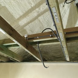 ANdom Instalacje Elektryczne - Montaż instalacji elektrycznej w nowo ocieplonym poddaszu, widoczne kable w peszlach przymocowane do drewnianych belek pokrytych metalowymi uchwytami, ocieplenie pianką poliuretanową.