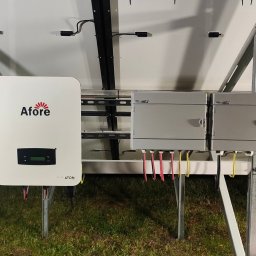 ANdom Instalacje Elektryczne - Widok od dołu na zamontowaną instalację fotowoltaiczną z widocznym inwerterem marki Afore i skrzynkami przyłączeniowymi, ustawioną na metalowej konstrukcji na tle trawy.