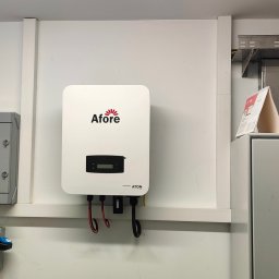 ANdom Instalacje Elektryczne - Biały falownik Afore zamontowany na ścianie w pomieszczeniu technicznym, obok szara skrzynka elektryczna i gniazdo siłowe. Widoczne kable zasilające. W tle metalowe korytka kablowe.