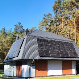 ANdom Instalacje Elektryczne - Dom z panelami fotowoltaicznymi na dachu z blachy trapezowej, w tle wysokie drzewa i błękitne niebo.