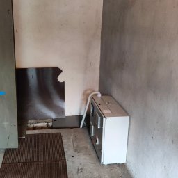 ANdom Instalacje Elektryczne - Wnętrze pomieszczenia z szarą, betonową ścianą, w którym znajduje się stara szafa elektryczna z opisem 'biurowiec' i nowa, biała jednostka klimatyzacyjna z podłączonym przewodem. Widoczna podłoga...