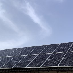 ANdom Instalacje Elektryczne - Panele słoneczne na dachu budynku jednorodzinnego, widoczne z dołu pod kątem, na tle błękitnego nieba z delikatnymi obłokami.