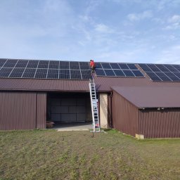 ANdom Instalacje Elektryczne - Montaż paneli słonecznych na dachu brązowego budynku gospodarczego; pracownik w czerwonej kurtce na drabinie, jasne niebo w tle.