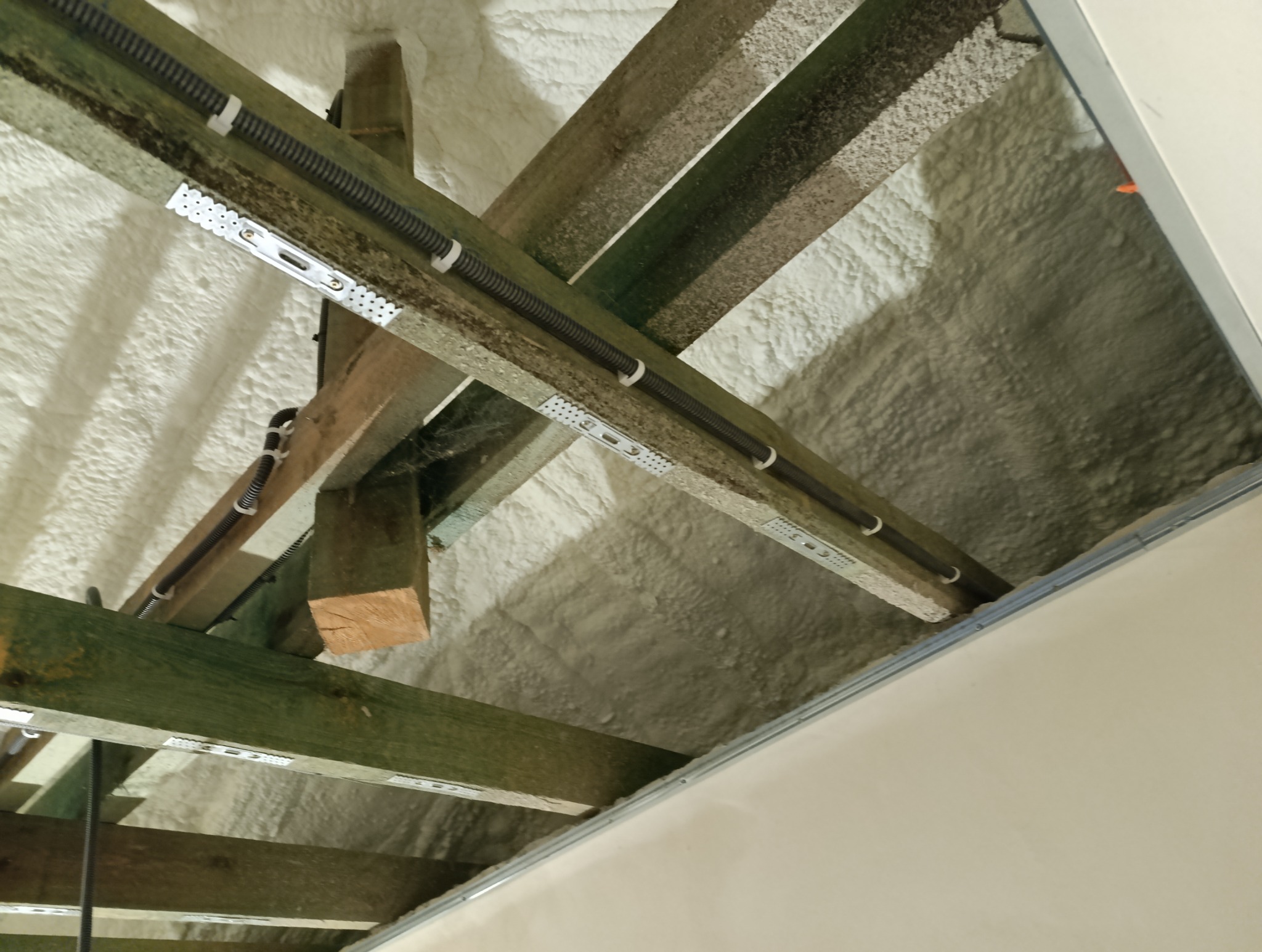 Izolacja poddasza pianą PUR, widoczne drewniane krokwie z przymocowanymi przewodami elektrycznymi w peszlach, metalowe łączniki krokwi.