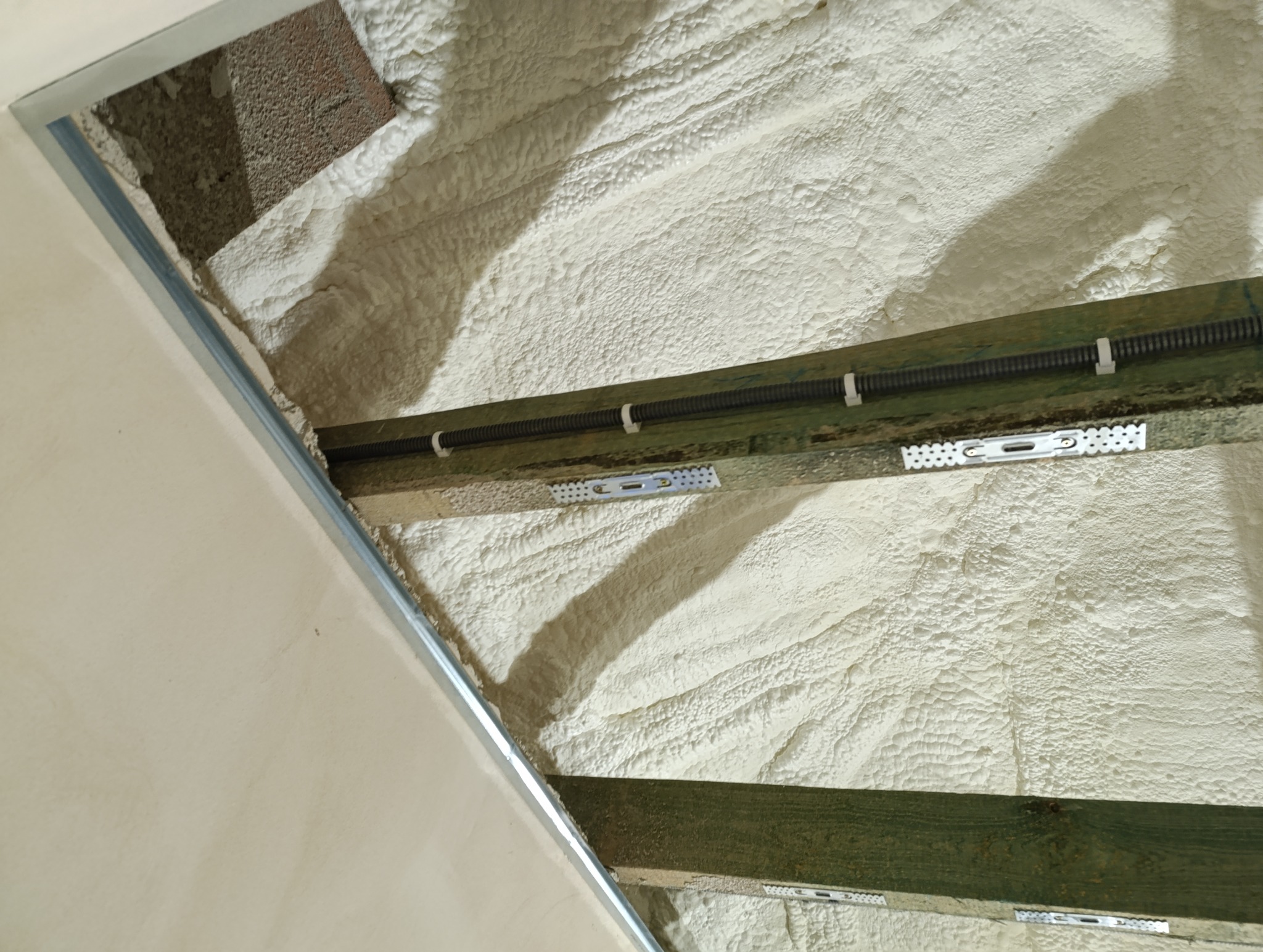 Poddasze ocieplone pianą poliuretanową, widoczne drewniane belki stropowe z przymocowaną peszlem elektryczną oraz fragment metalowego profilu sufitu podwieszanego.