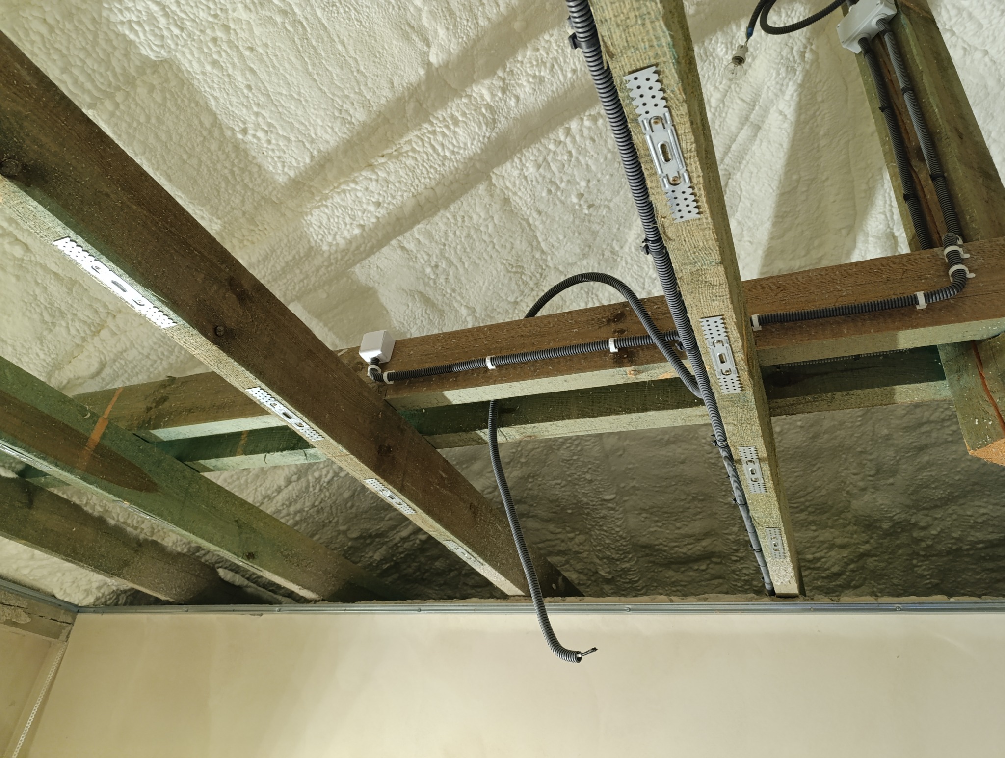 Montaż instalacji elektrycznej w nowo ocieplonym poddaszu, widoczne kable w peszlach przymocowane do drewnianych belek pokrytych metalowymi uchwytami, ocieplenie pianką poliuretanową.