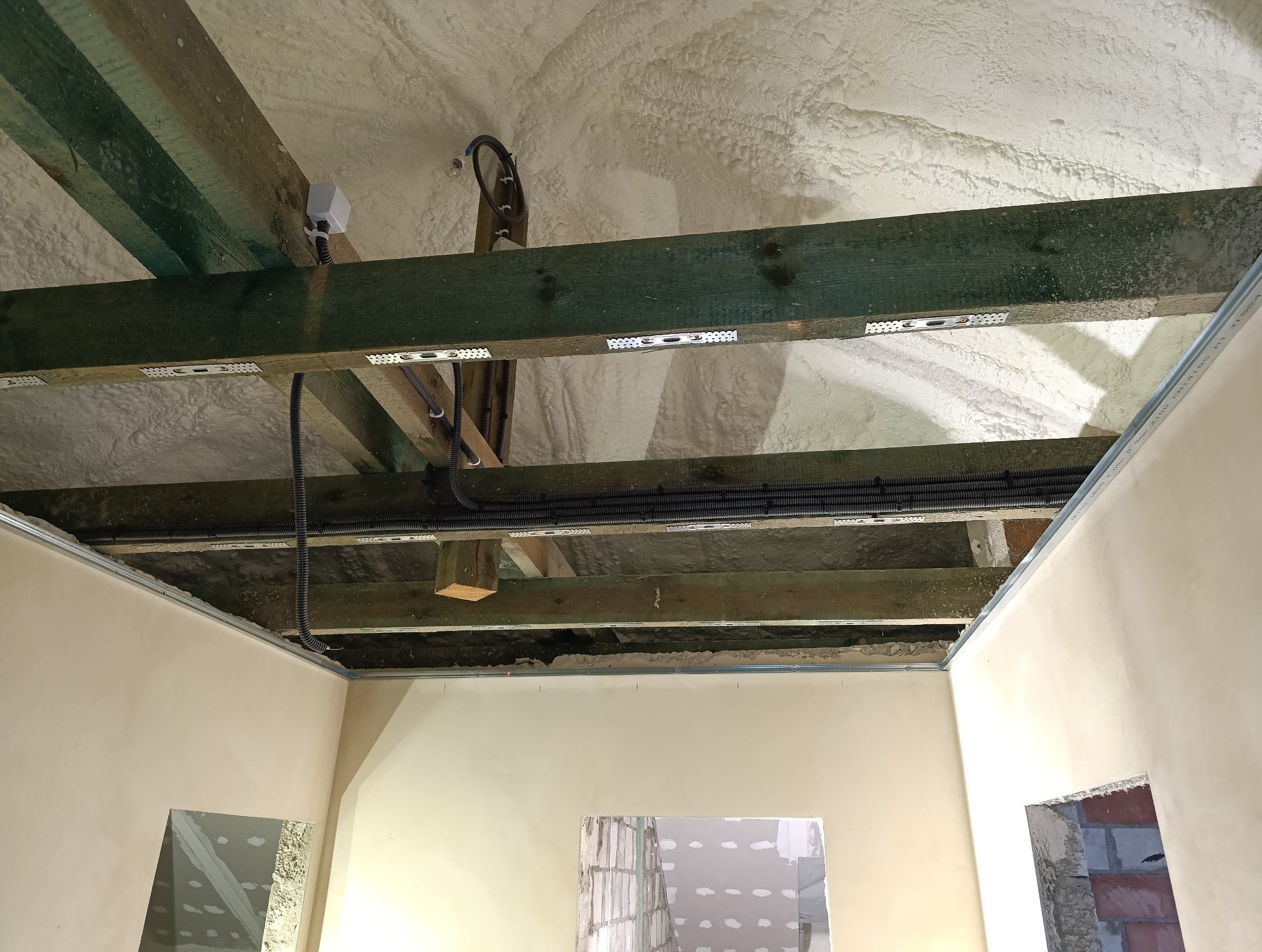 Izolacja pianą PUR poddasza z widocznymi belkami stropowymi, przewodami elektrycznymi w peszlach i profilem sufitu podwieszanego.