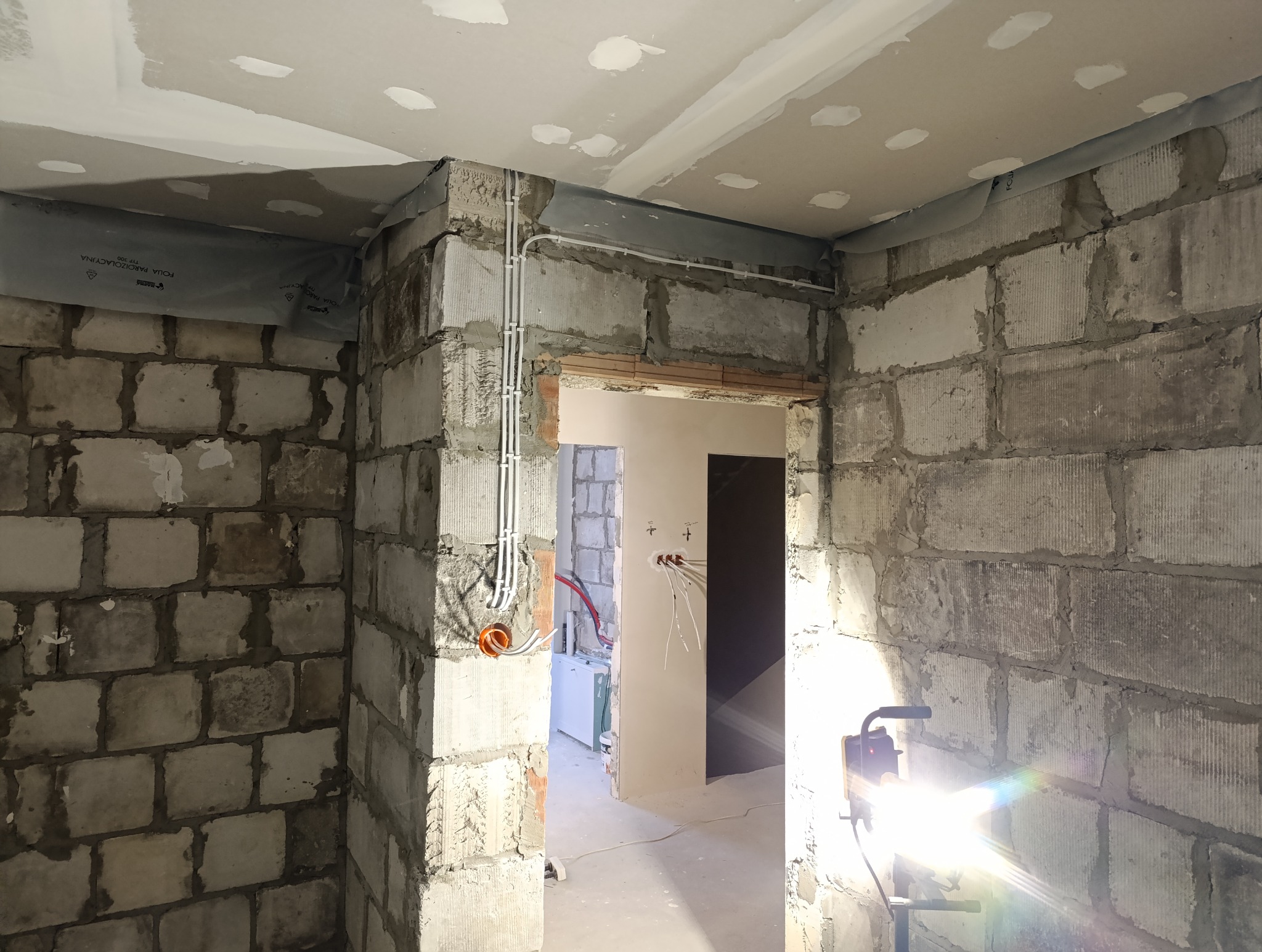 Surowe wnętrze w trakcie remontu: widoczne kable elektryczne prowadzone wzdłuż ściany z betonu komórkowego, instalacja elektryczna w puszce, sufit z płyt gipsowo-kartonowych z widocznymi łączeniami...