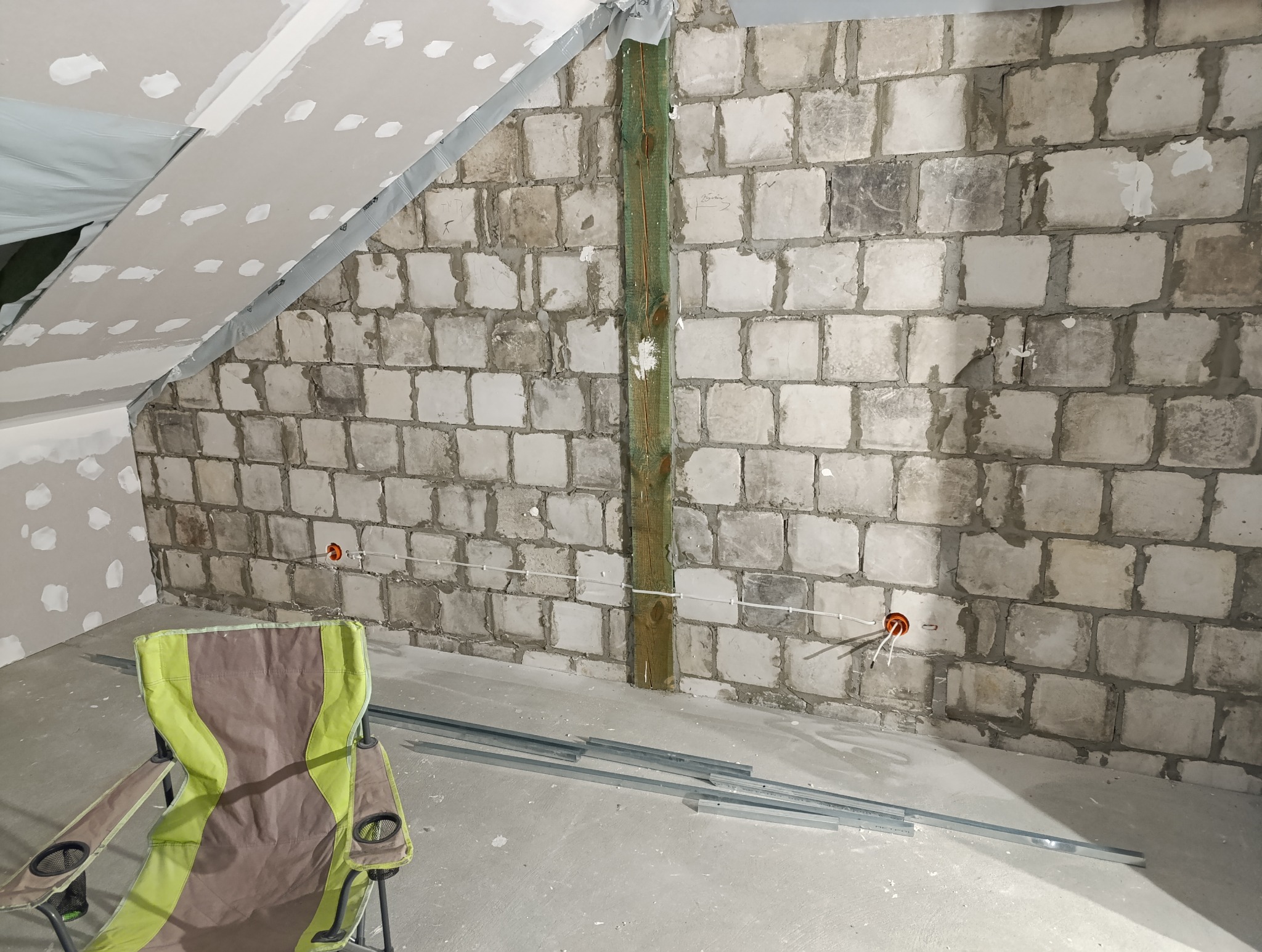 Adaptacja poddasza: ściana z białej cegły, widoczne przewody elektryczne w pomarańczowej puszce, częściowo zamontowane płyty gipsowo-kartonowe na skosach, drewniany słup wzmacniający, profil...