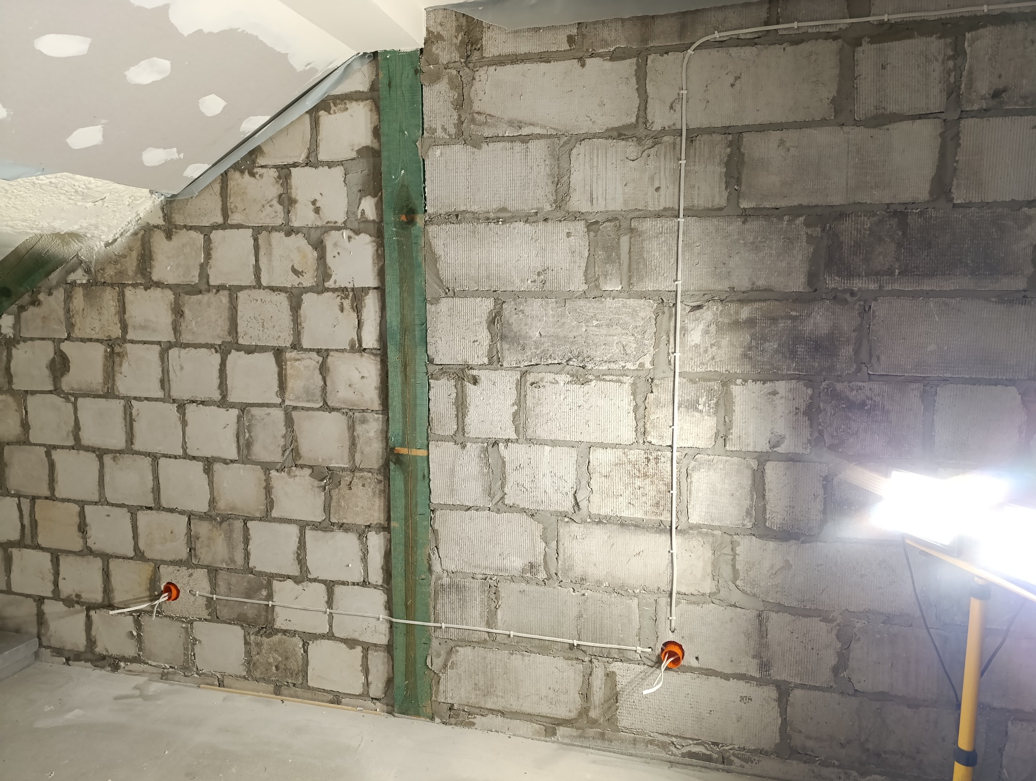 Ściana z betonu komórkowego w trakcie prac elektrycznych, widoczne kable w peszlach, puszki instalacyjne oraz drewniana listwa pionowa, oświetlona lampą roboczą.