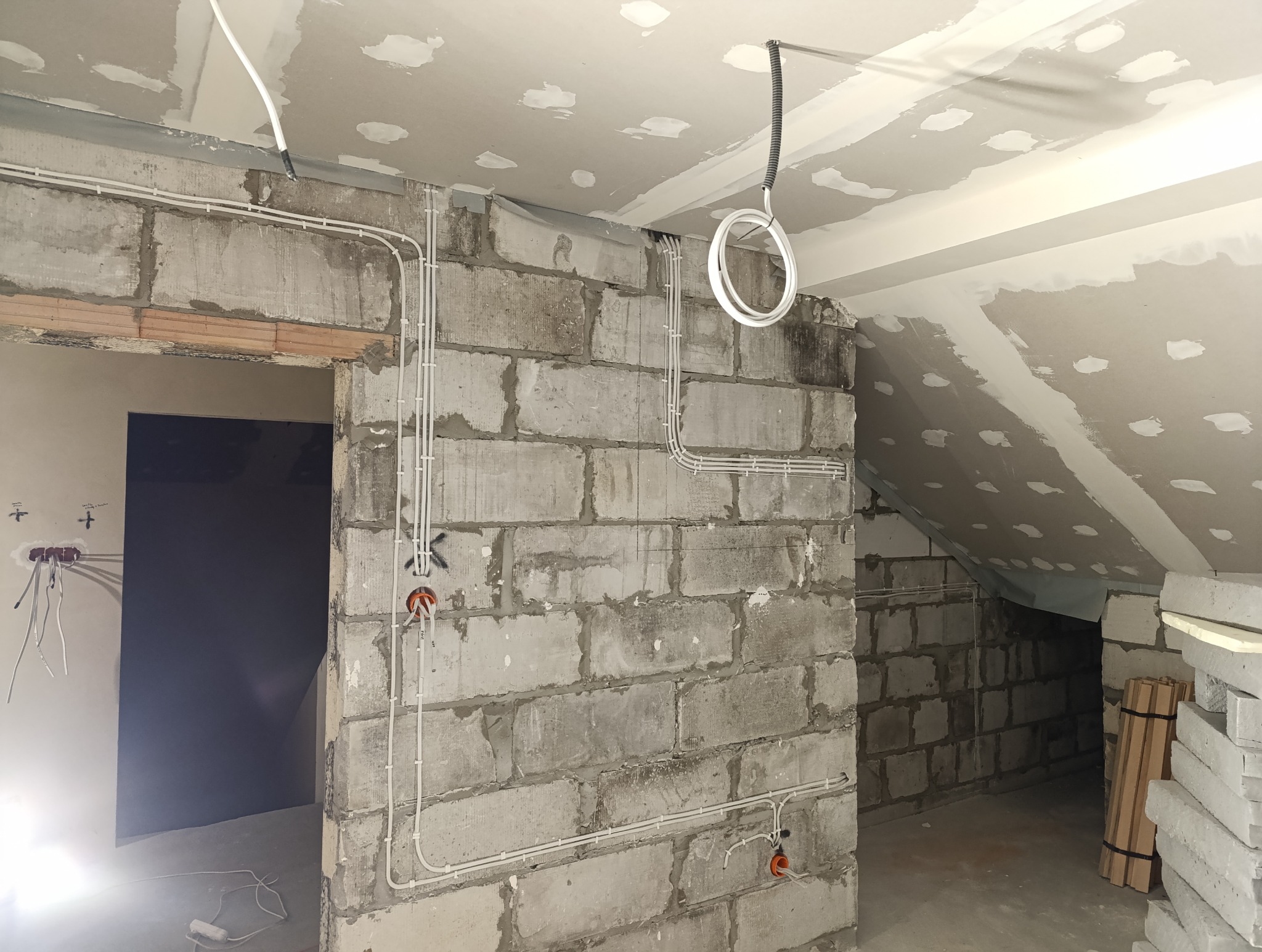 Instalacja elektryczna w surowym wnętrzu: białe przewody prowadzone po ścianach z betonu komórkowego, widoczne puszki instalacyjne i częściowo wykończony sufit poddasza z widocznymi łatami gipsowymi.