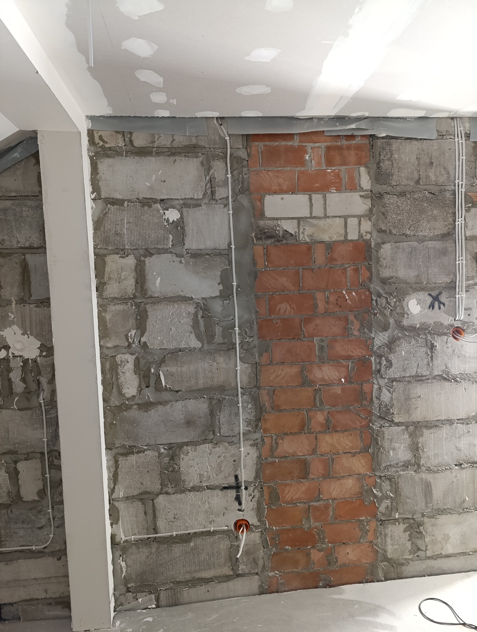 Wnętrze w trakcie remontu: surowa ściana z betonu komórkowego i czerwonej cegły, widoczne przewody elektryczne w peszlach i puszki instalacyjne, sufit z płyt kartonowo-gipsowych w trakcie...